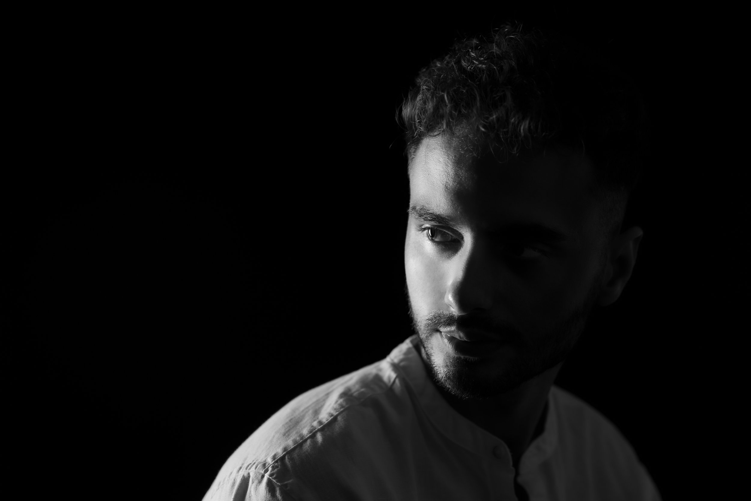 Black and white portrait of a young man with curly hair, looking down, with shadows on one side of his face against a dark background.