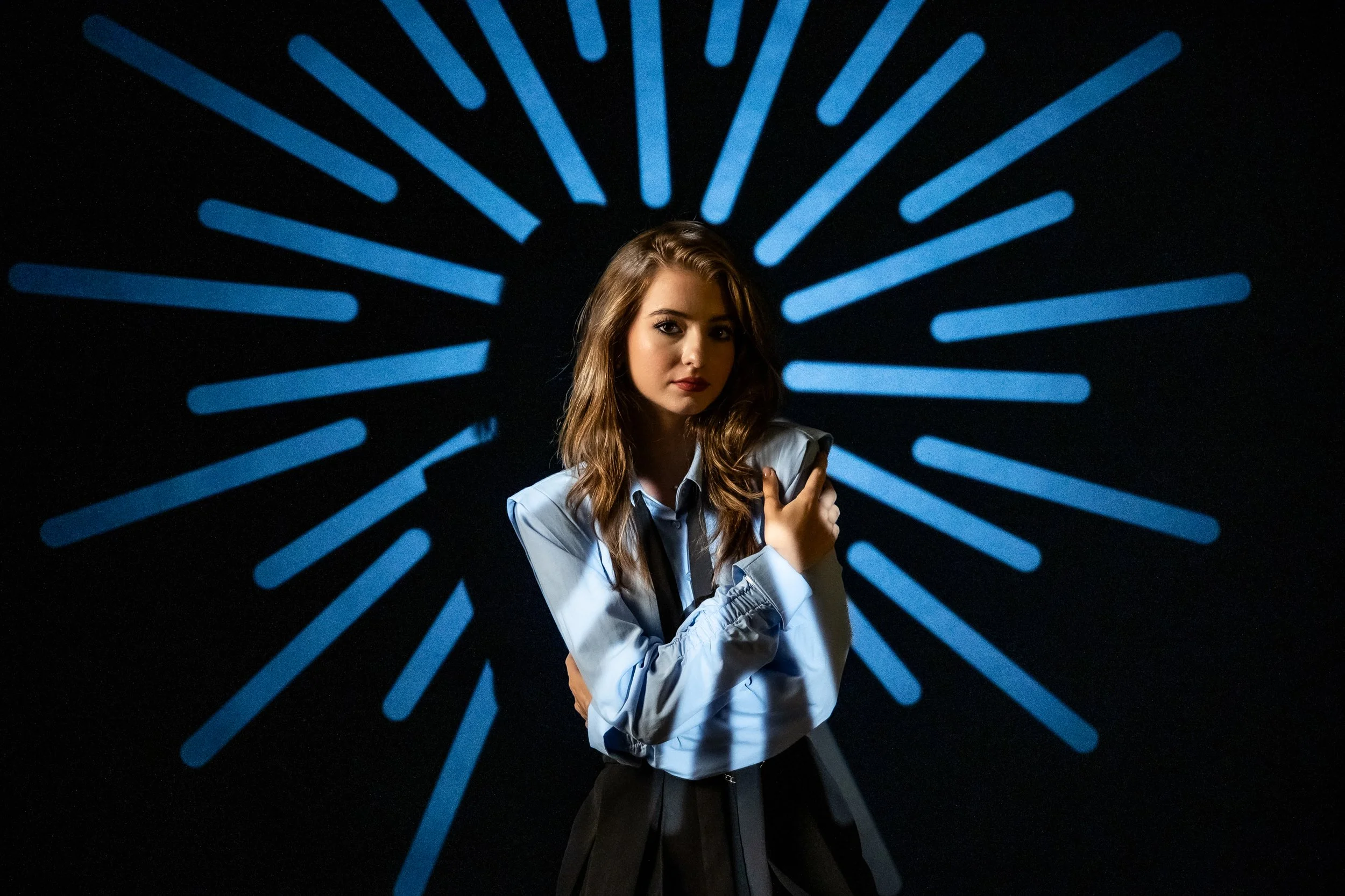 A young woman with wavy brown hair, wearing a light blue button-up shirt with rolled-up sleeves, stands with her arms crossed against a black background with radiating blue lines.