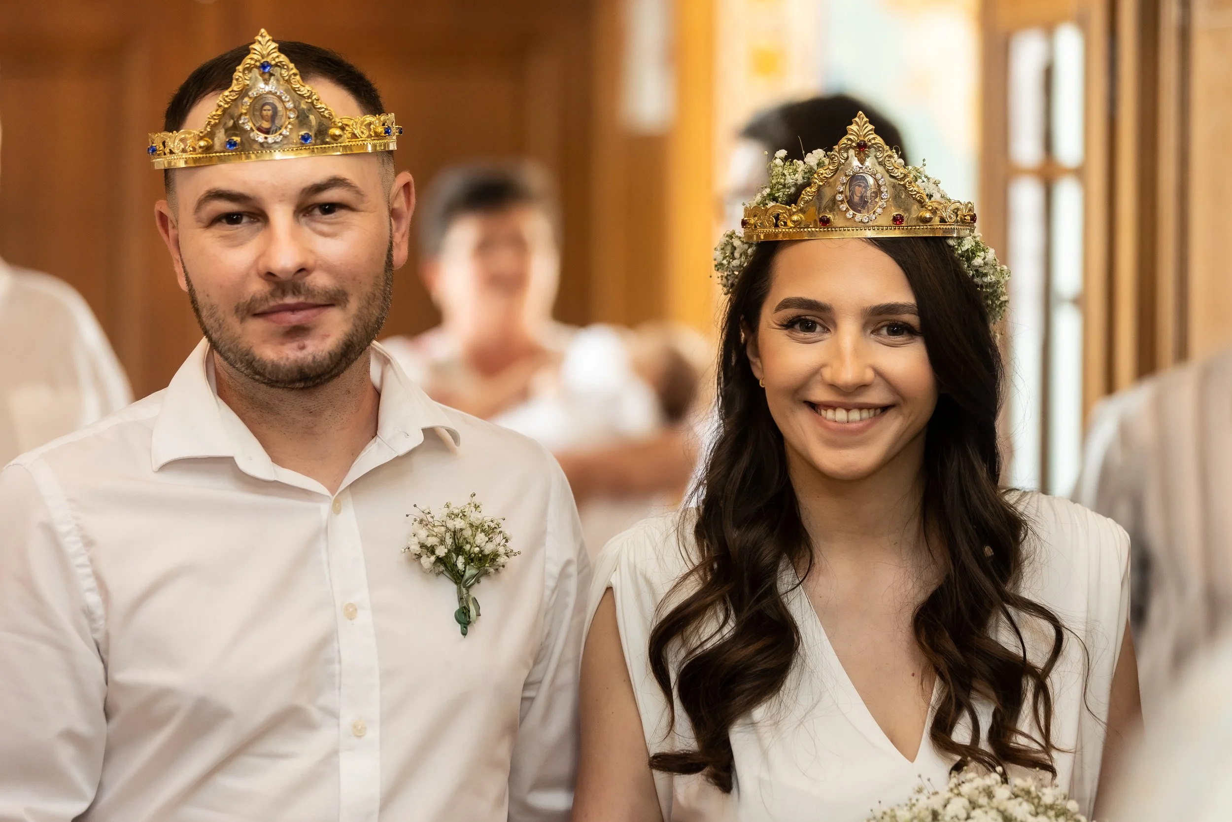 A smiling couple wearing wedding crowns, standing next to each other during a wedding ceremony, dressed in white. The man has a short beard and is holding a small bouquet, while the woman has long dark hair and is also holding a small bouquet.