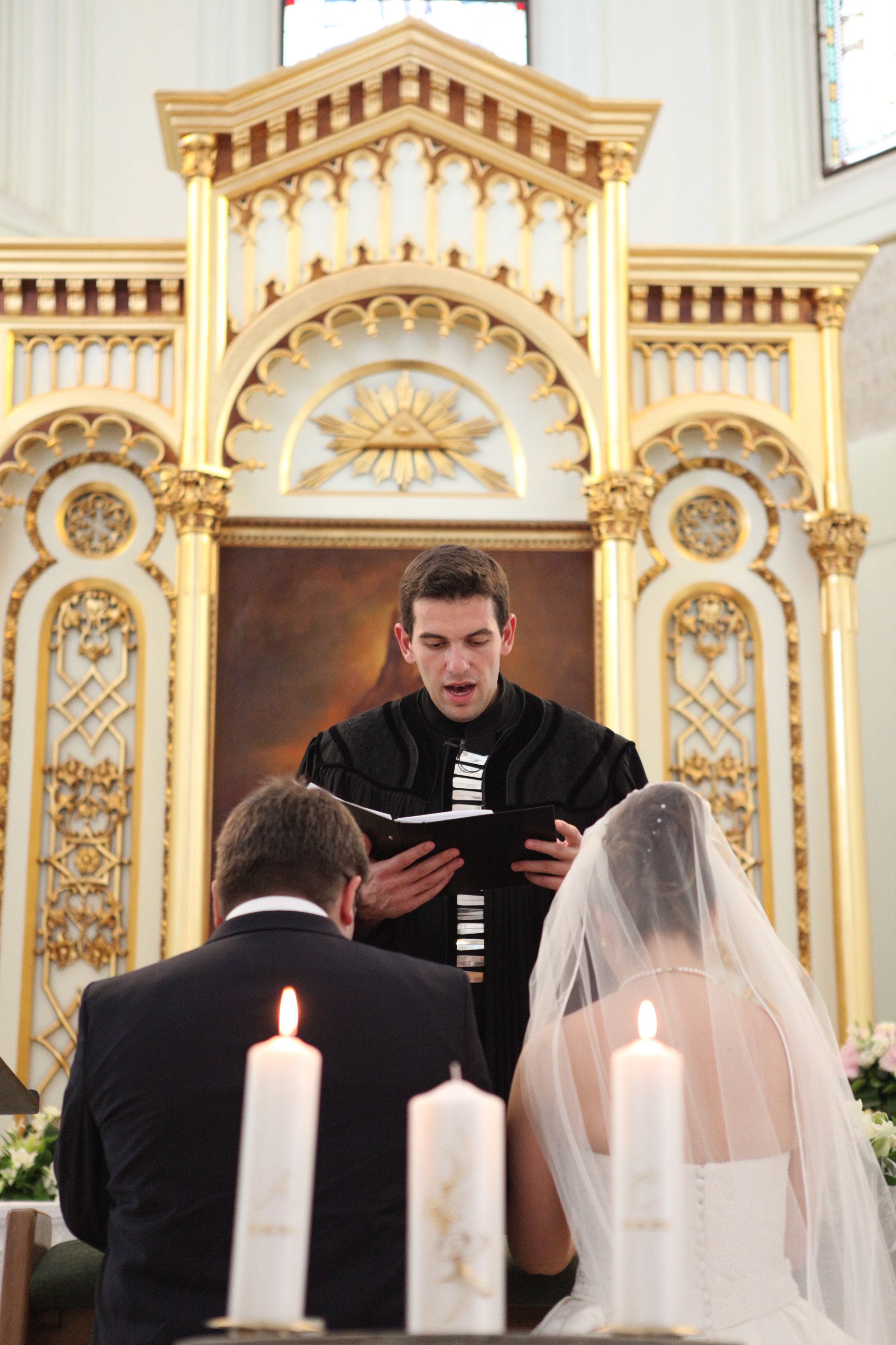 A groom and bride kneeling at the altar during a wedding ceremony in a church. The officiant stands behind them reading from a book. Candles are in the foreground, and an ornate gold and white altarpiece with religious symbols is behind the officiant