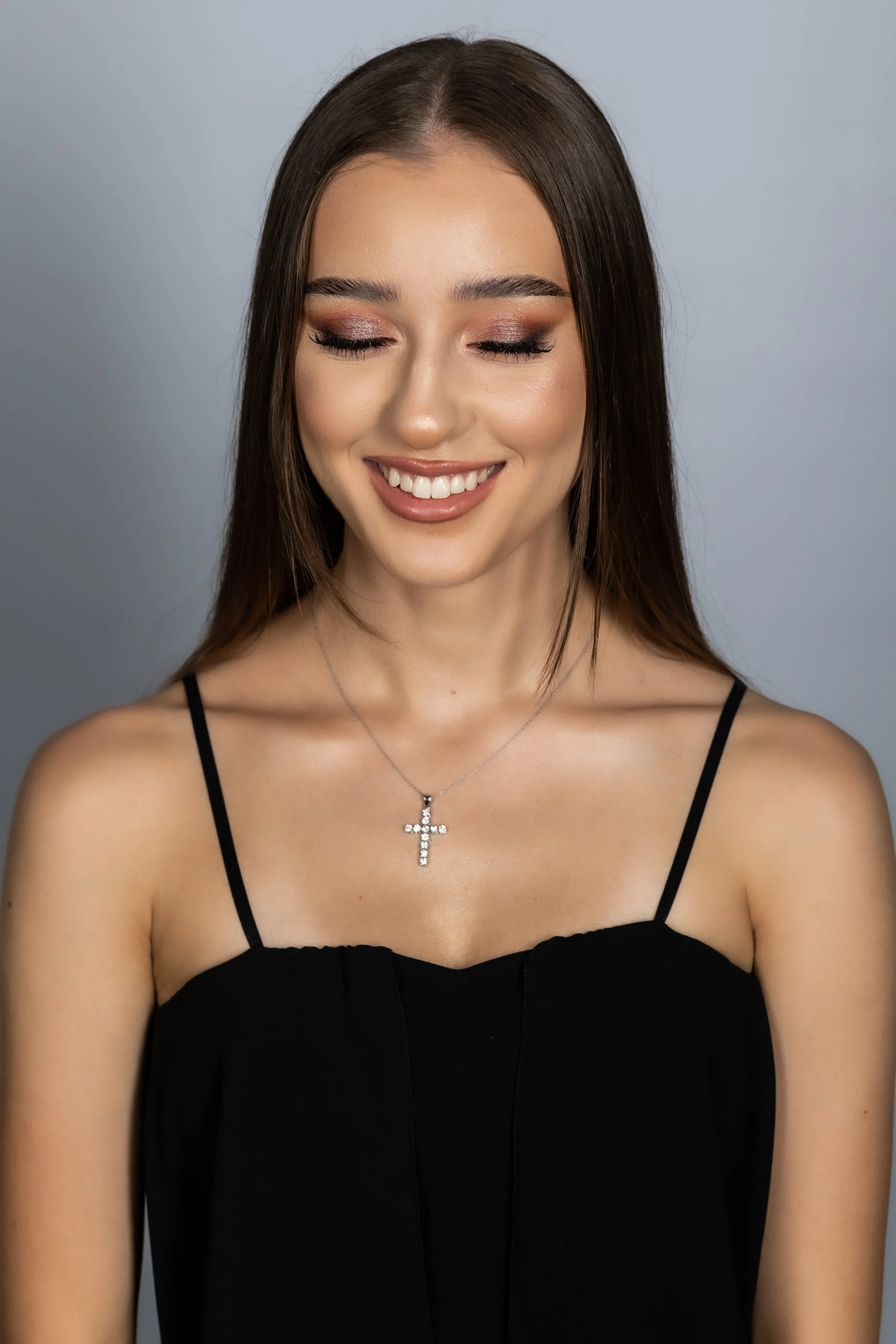 Portrait of a young woman with long brown hair, closed eyes, and a smile. She is wearing a black spaghetti strap top and a silver cross necklace. The background is plain and light-colored.