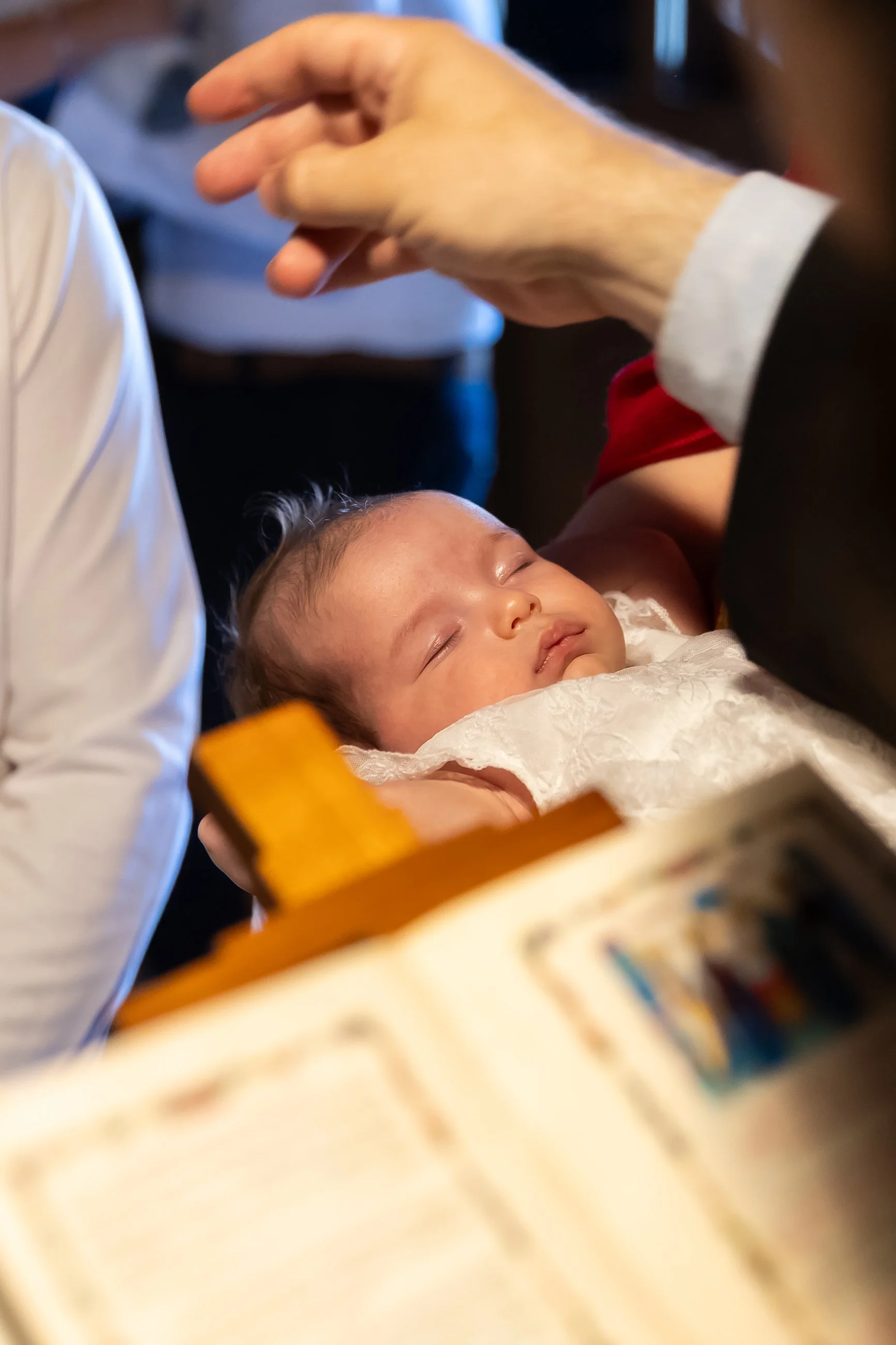 Close-up of a sleeping baby with closed eyes, lying on a white blanket, being held gently by adult hands, in a cozy indoor setting.