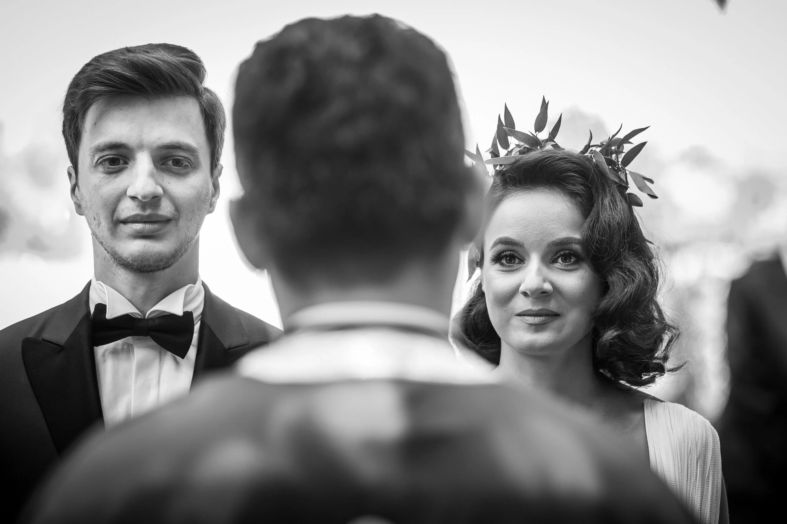 A black and white photo of a wedding ceremony showing a groom in a tuxedo with a bow tie and a bride with curly hair and a laurel crown, facing a person with short hair, possibly an officiant.