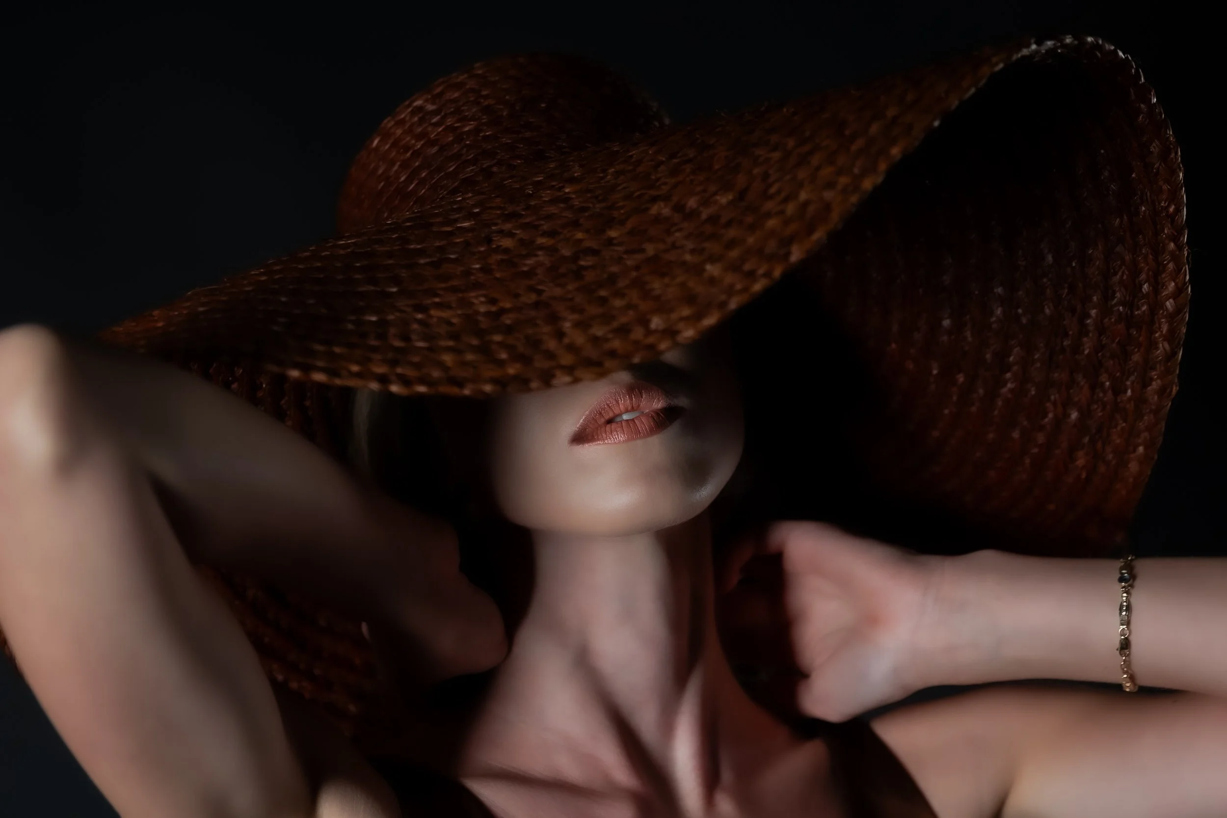 A woman with a wide-brimmed brown hat covering part of her face, showing her lips and one eye, with skin illuminated against a black background.