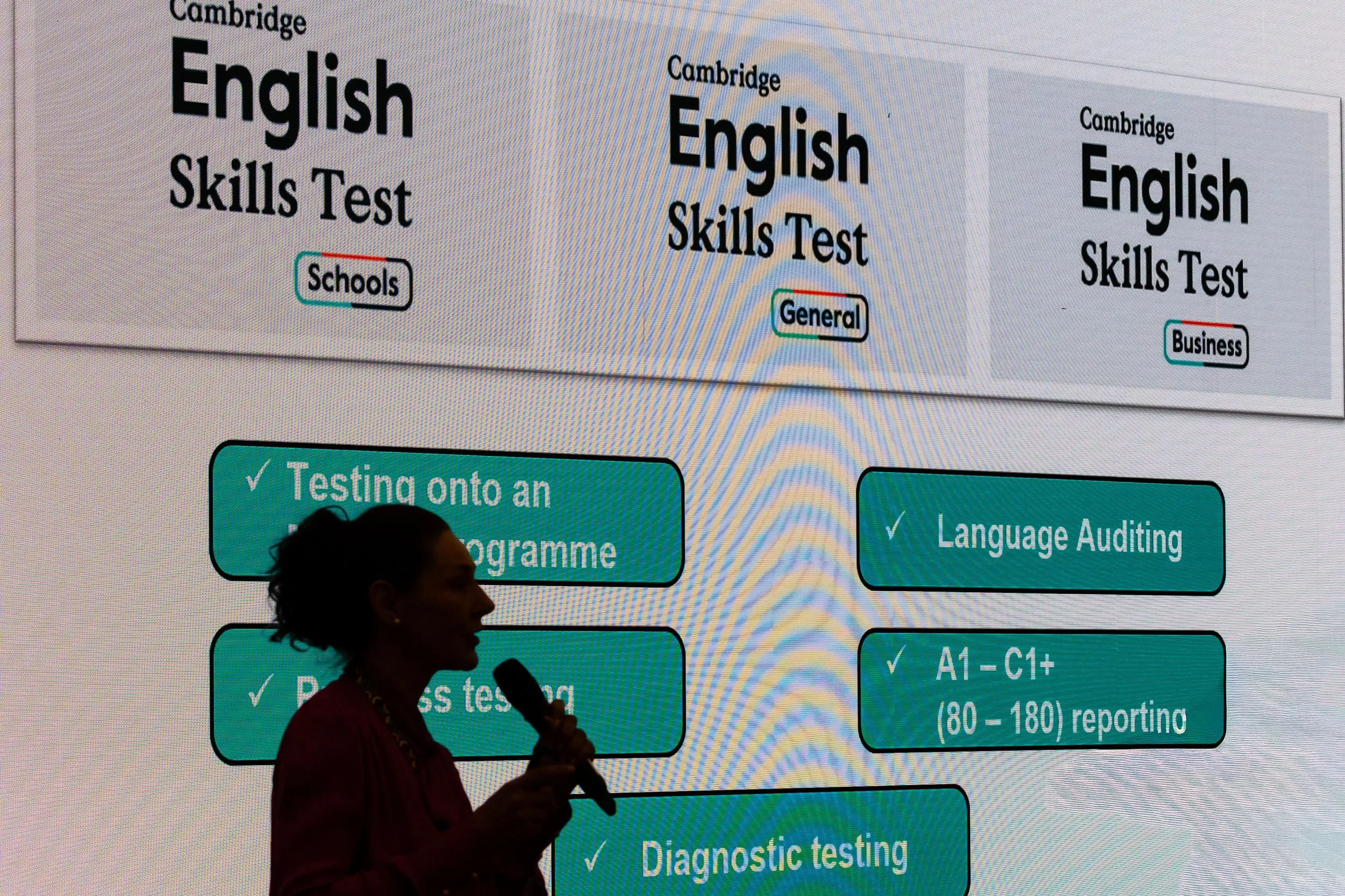 A person holding a microphone stands in front of a large presentation screen displaying information about the Cambridge English Skills Test.