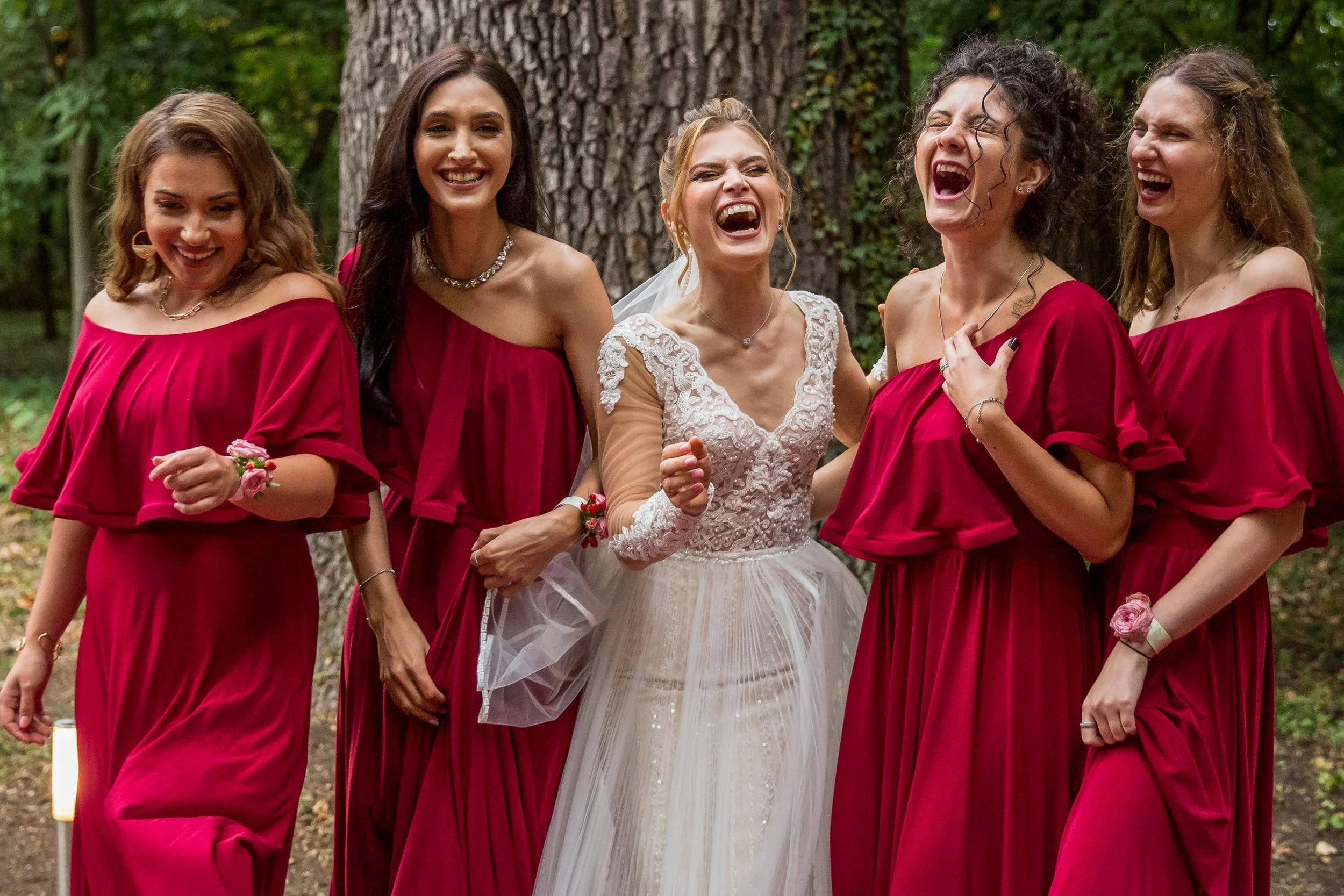 A bride and her bridesmaids celebrating outdoors in front of a large tree. The bride is in a white wedding dress, and the bridesmaids are in matching red dresses. They are all laughing and smiling.