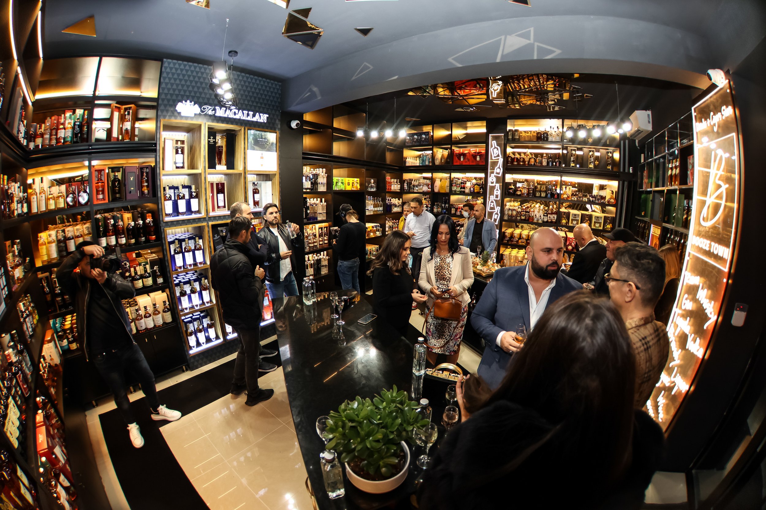 People gathered in a modern store with shelves of liquor bottles, some engaging in conversation, others browsing or taking photos, with a black and gold interior design.