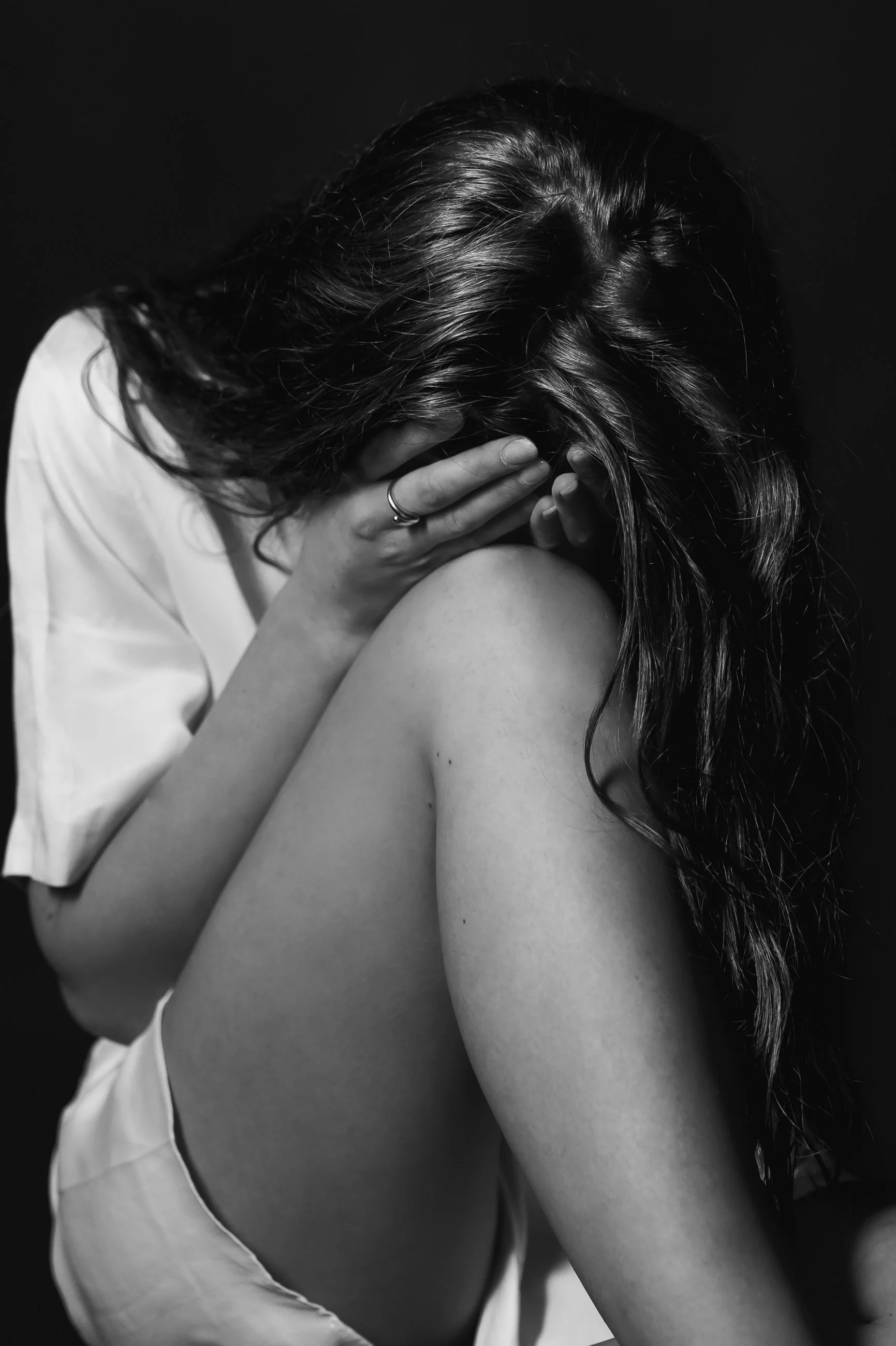 A person with long, wavy hair covering their face, holding their head in one hand and resting their arm on their knee, against a dark background.