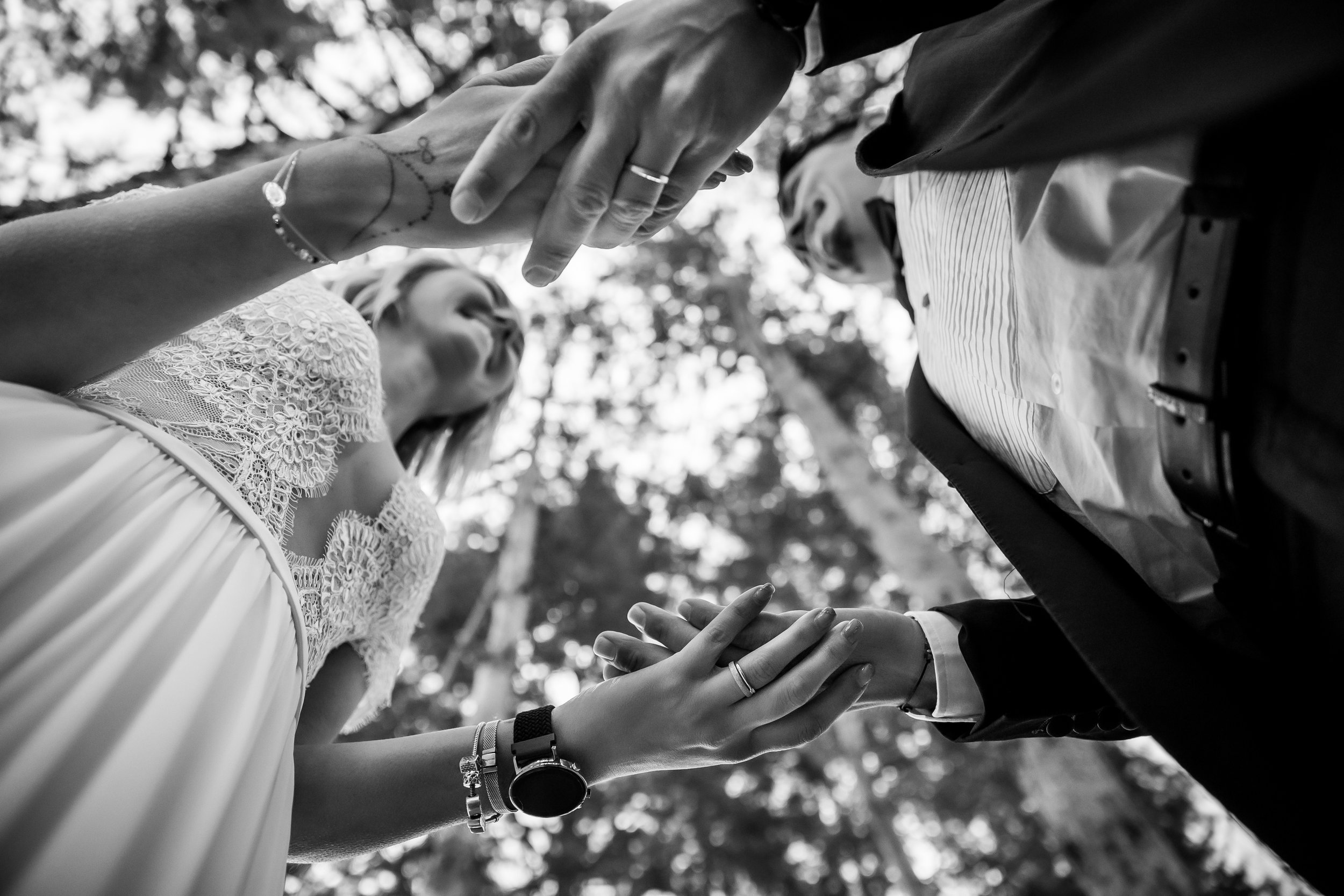 Black and white photo of a couple holding hands during a wedding ceremony, with trees and sunlight in the background.