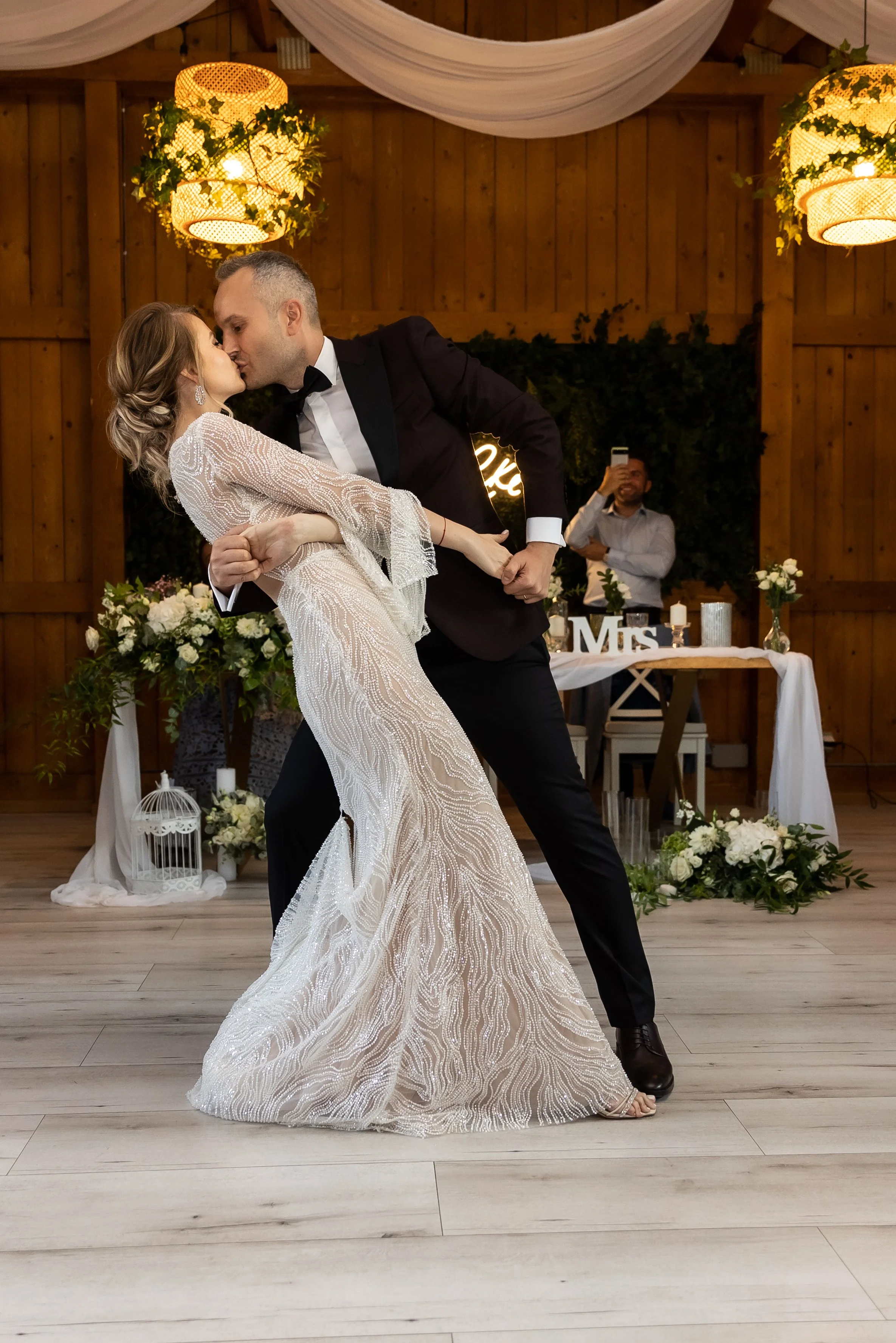 A bride and groom sharing a kiss and dance at their wedding reception in a decorated rustic venue, with floral arrangements and a man taking a photo in the background.