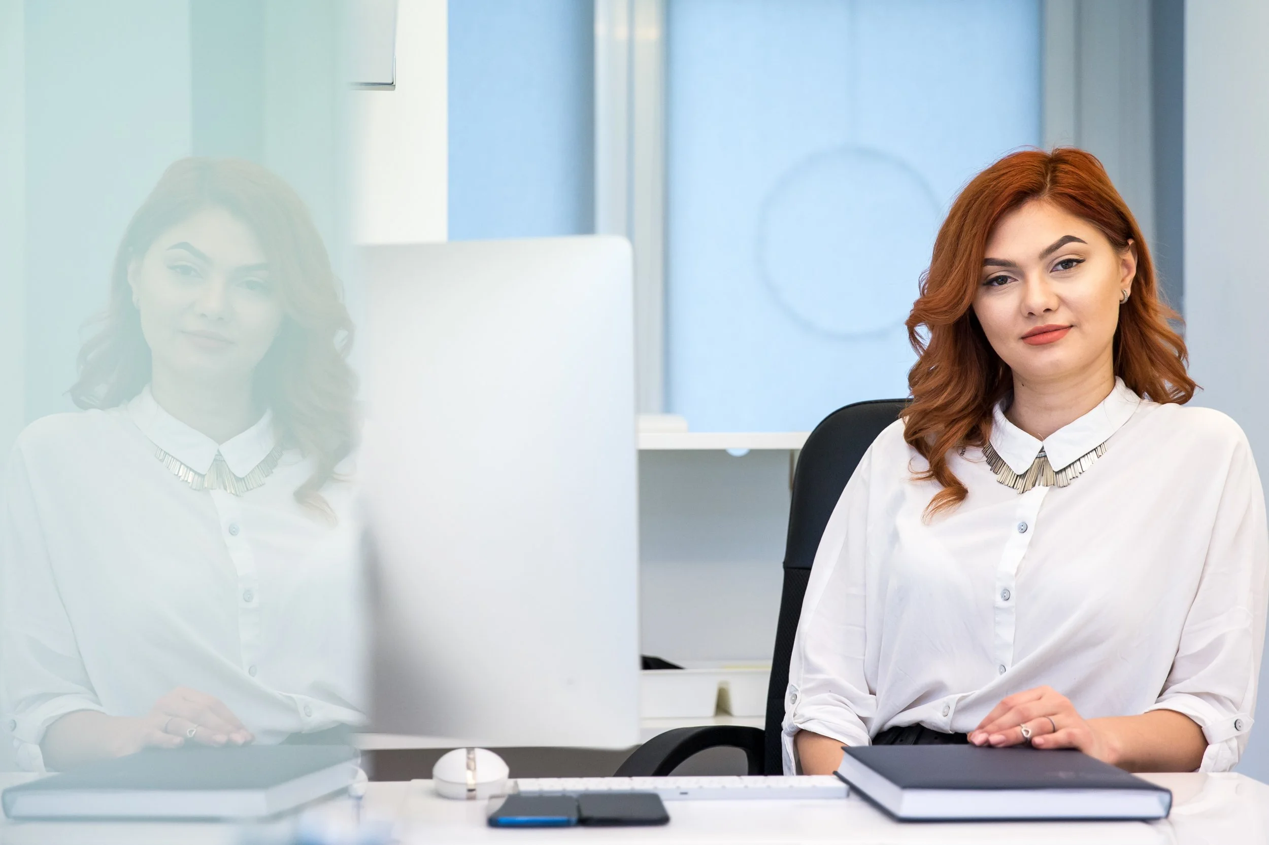 A woman with red hair in a white blouse sitting at a desk with a printer and papers, looking at the camera, with her reflection visible on a glass partition.