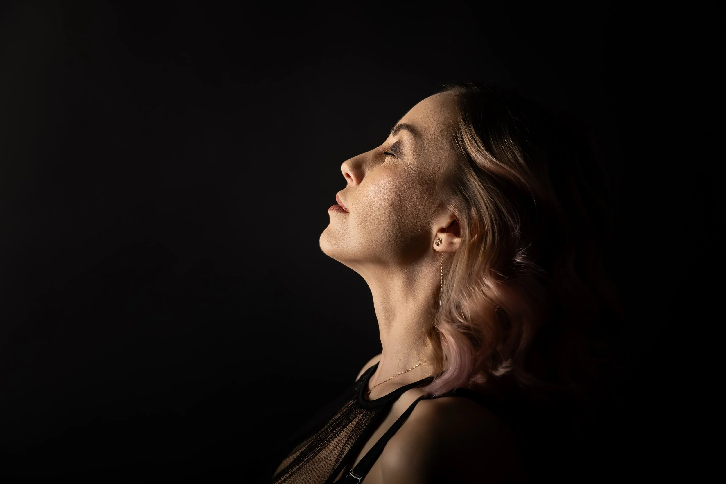 Side profile of a woman with short, wavy, light brown hair with pink tips, eyes closed, wearing earrings and black lace clothing against a black background.