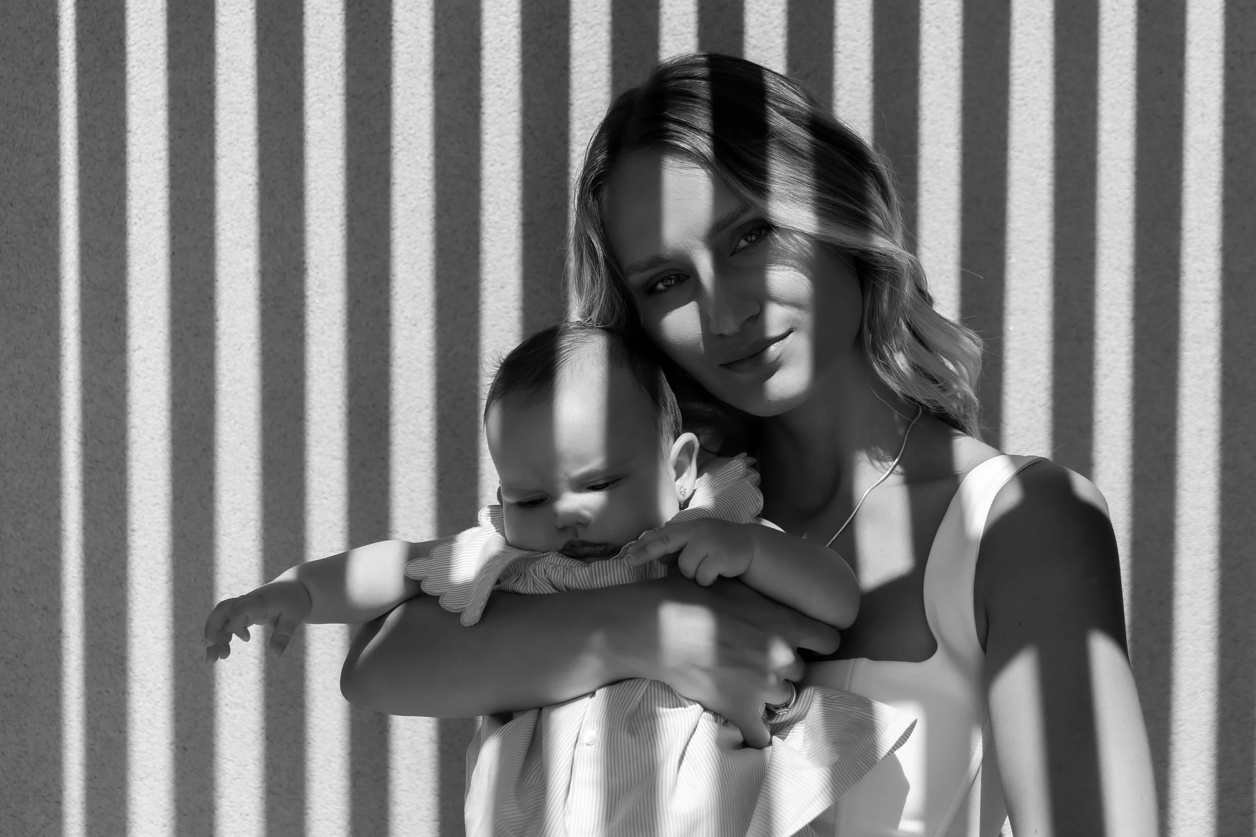 A woman with wavy hair holding a young child in her arms, both looking at the camera, with shadows of vertical lines cast across them on a textured wall.