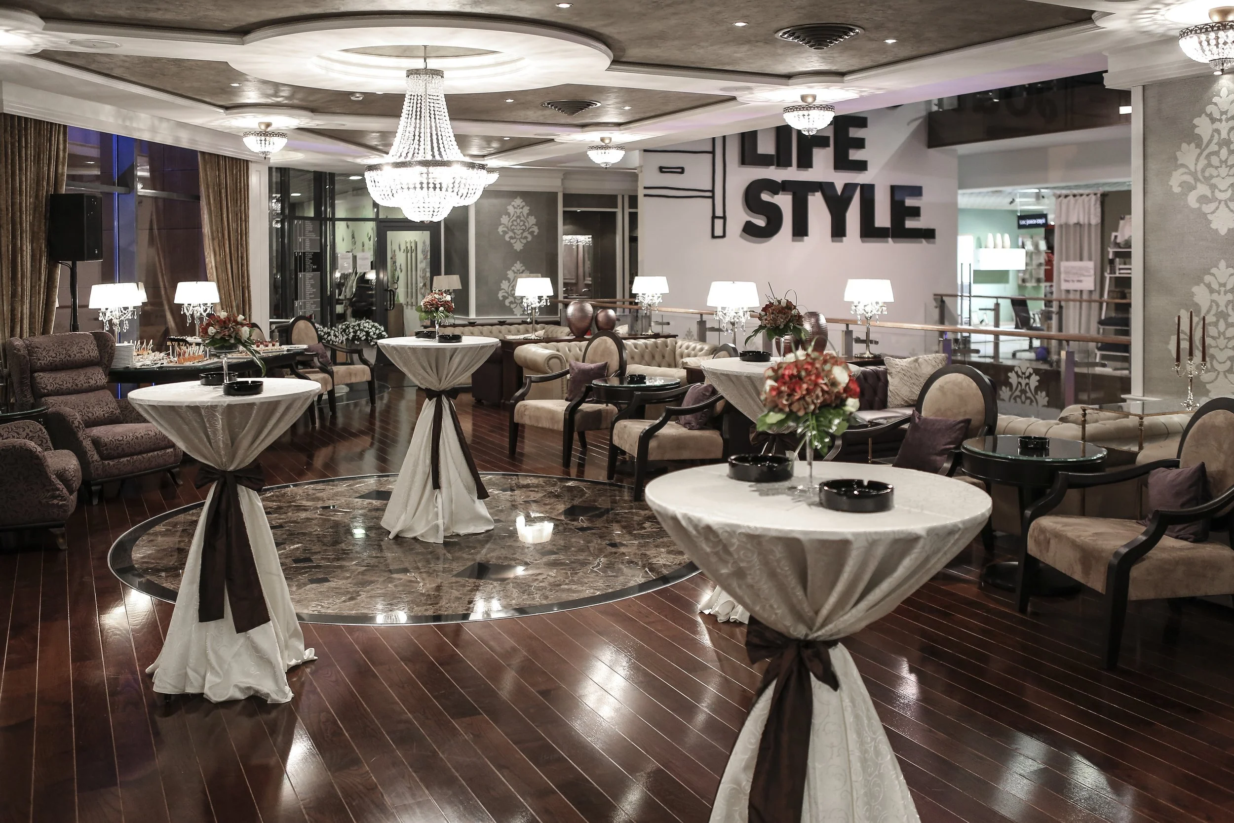 Elegant hotel lobby or lounge area with round tables covered with white tablecloths, surrounded by chairs and sofas. Decorative lamps and floral arrangements are on the tables. A large chandelier hangs from the ceiling, and a wall displays the words 