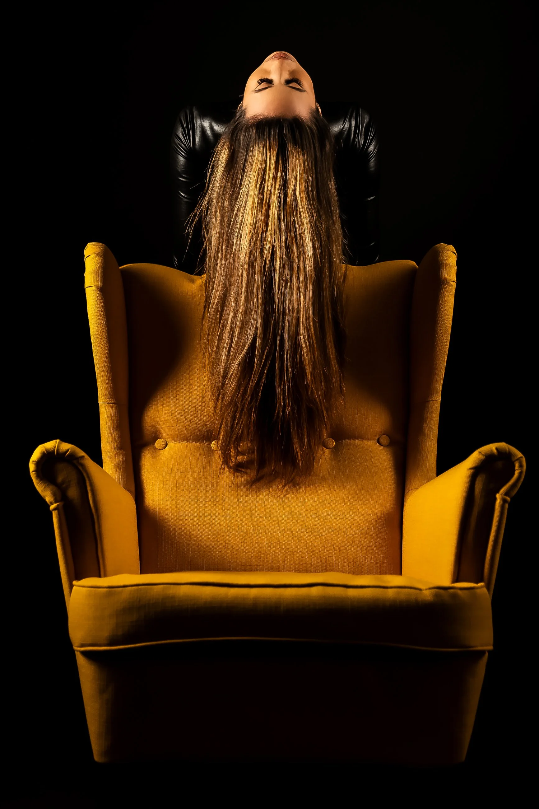A woman with long brown hair, eyes closed, leaning back on a yellow armchair against a black background.