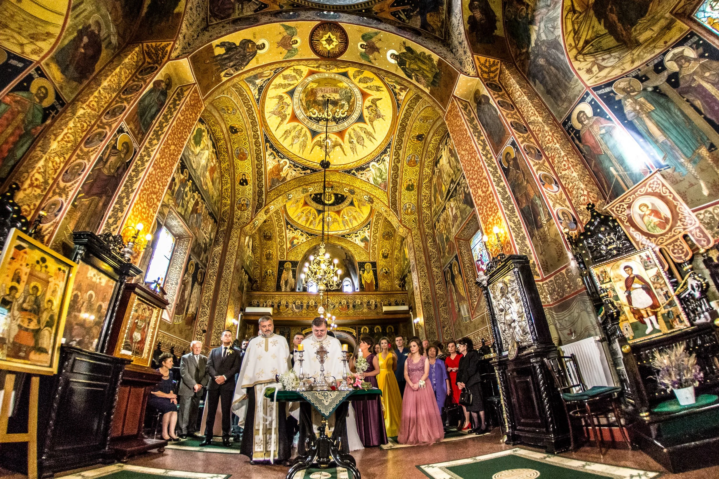 A religious ceremony taking place inside an ornate church with gold embellishments and religious iconography covering the walls and ceiling. Several clergy members are conducting the service while a congregation of people, dressed in formal attire, p