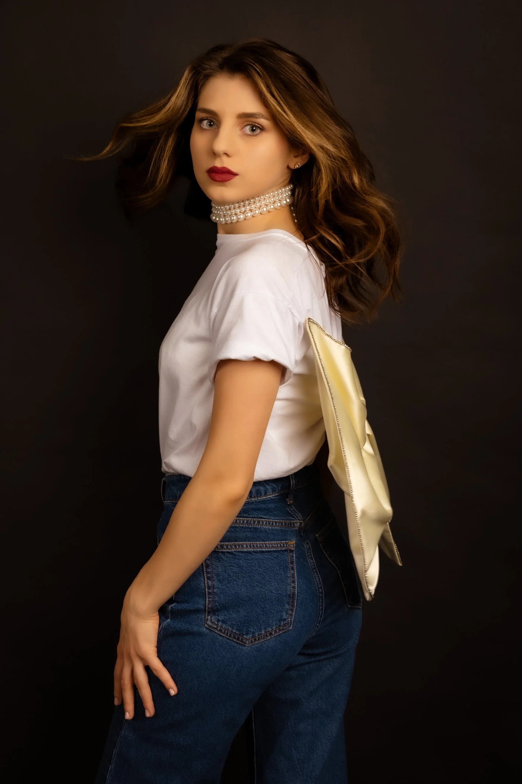 Young woman with wavy brown hair wearing a white t-shirt, dark blue jeans, a pearl choker, and holding a yellow clutch bag against a black background.