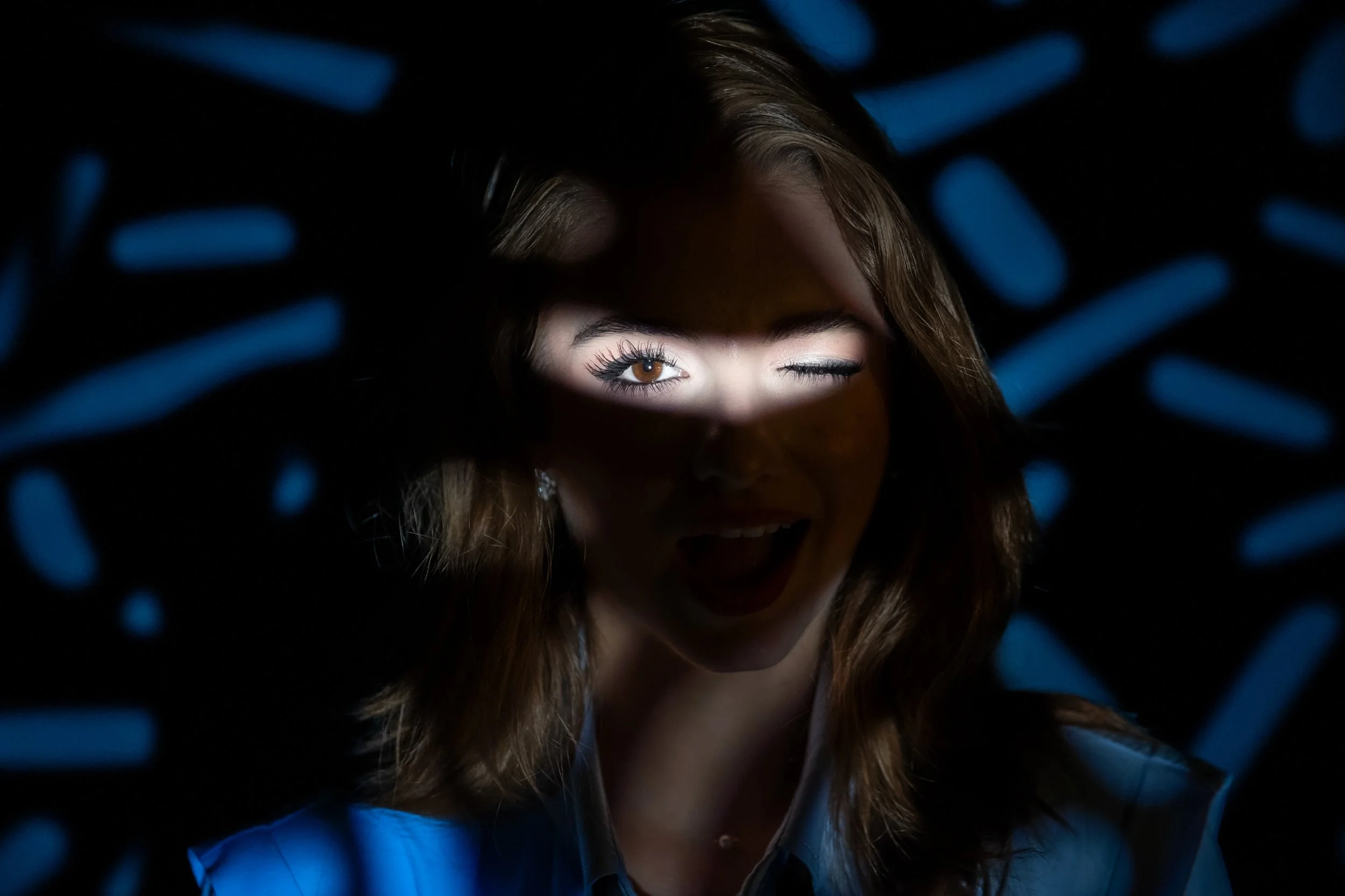 A woman with brown hair winking and smiling, illuminated by a circular pattern of blue light in a dark environment.