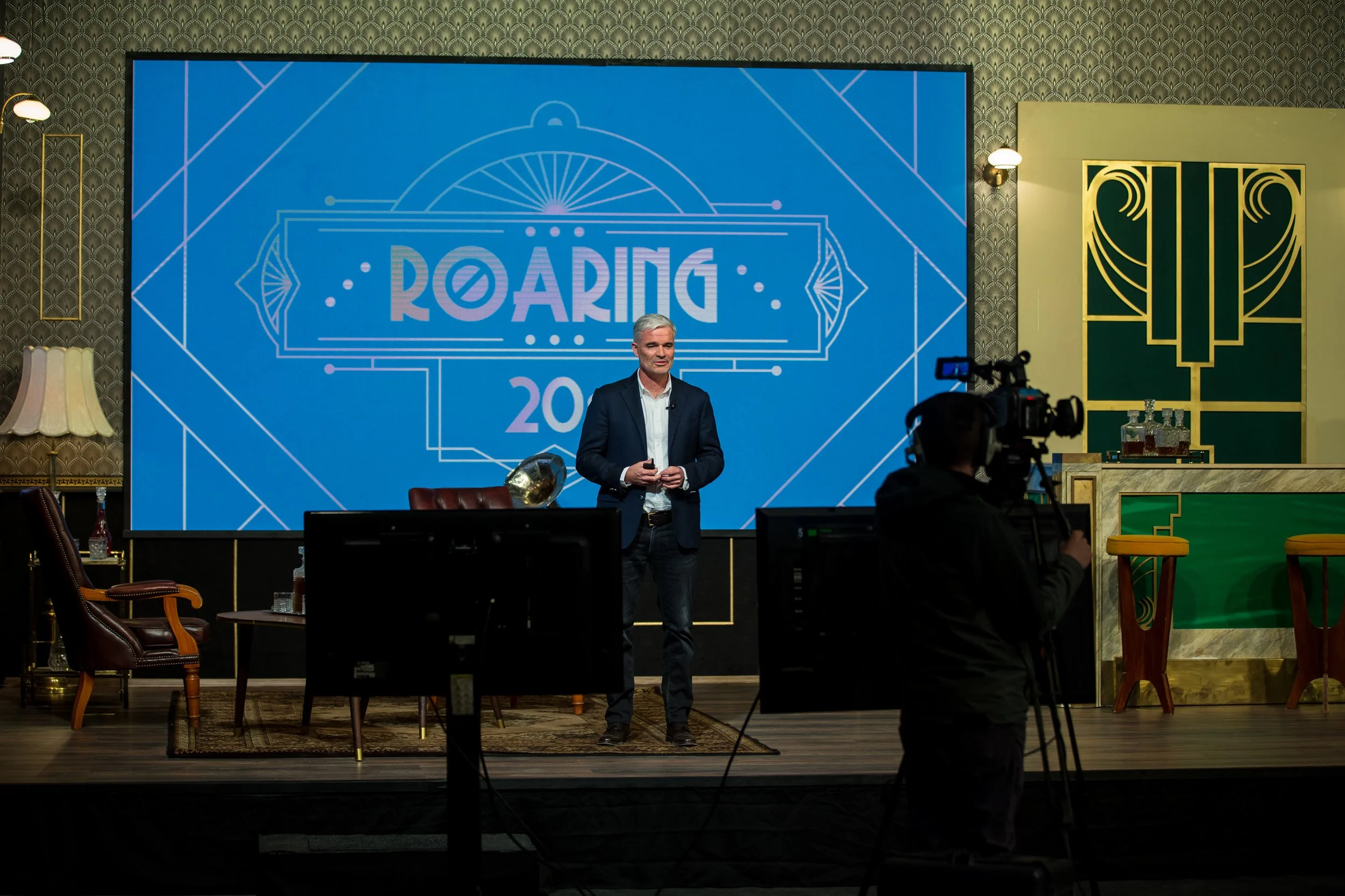 A man in a suit giving a presentation on stage at a conference or event, with a large screen behind him displaying the word 'ROARING' and other graphics, with cameras and audience members visible in the foreground.