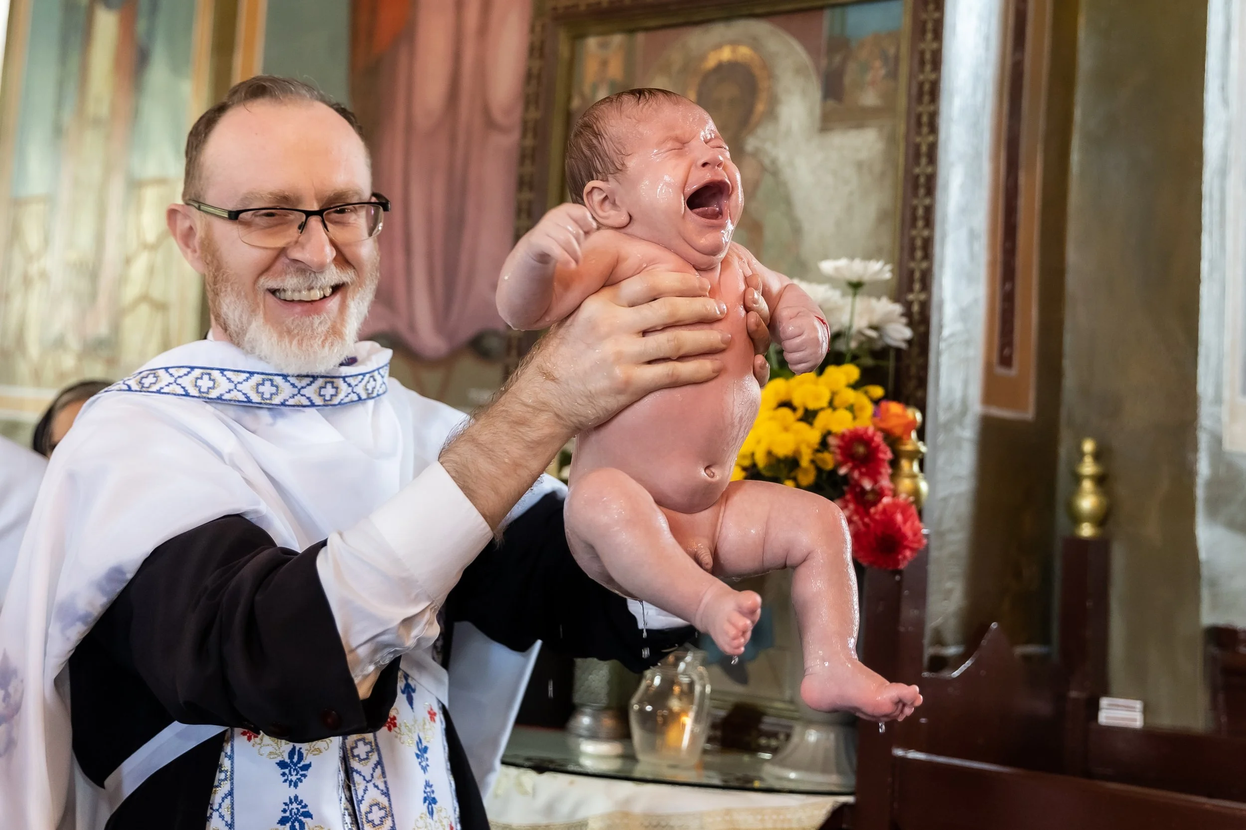 A priest holding a crying baby during a baptism ceremony inside a church with religious icons and flowers in the background.