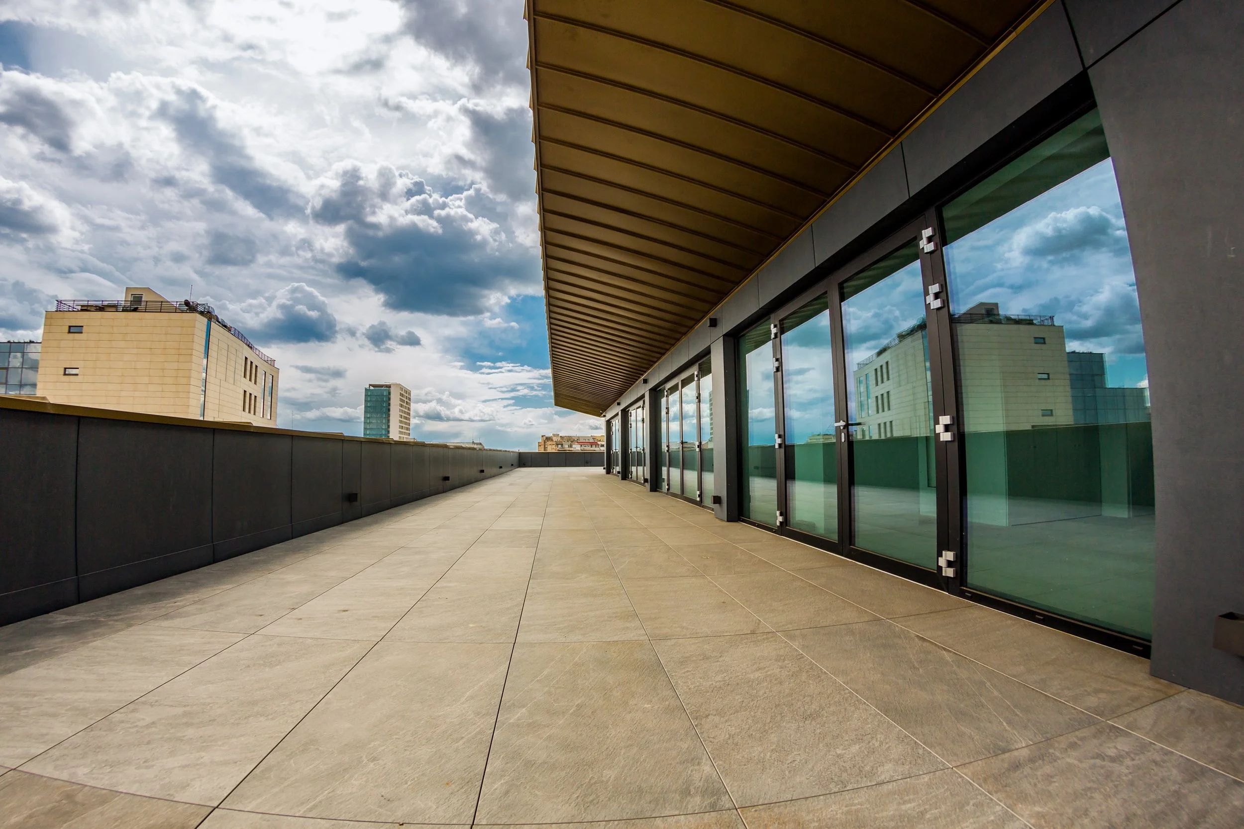 Empty outdoor terrace with large glass windows, modern beige buildings, and a partly cloudy sky.