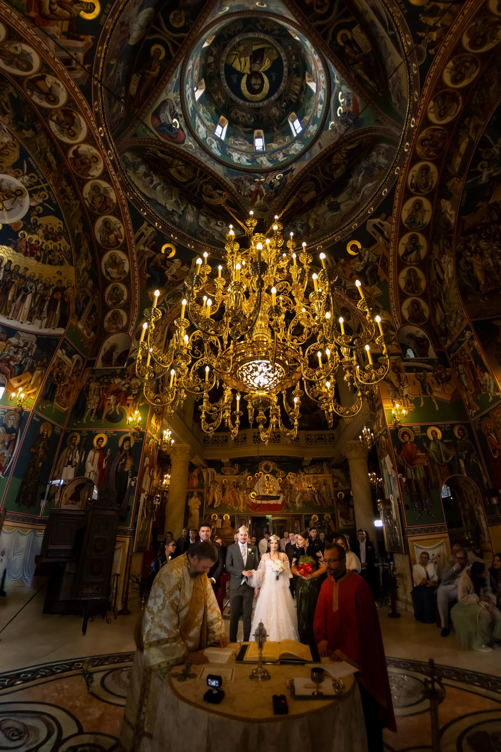 A wedding ceremony taking place inside a richly decorated Orthodox church with gold chandelier, detailed religious icons on the walls, a high decorated ceiling, and a congregation of people including the bride and groom standing at the altar.