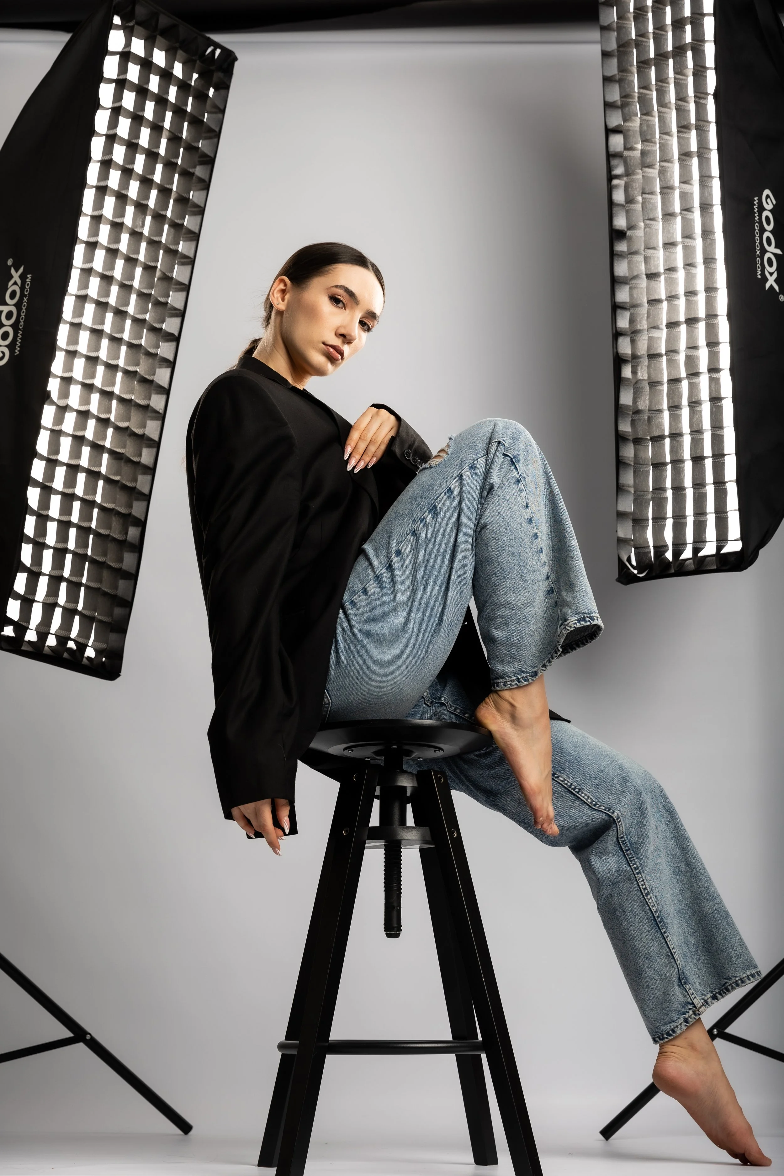 A young woman in a black blazer and jeans, sitting on a stool in a photography studio, surrounded by professional lighting equipment.