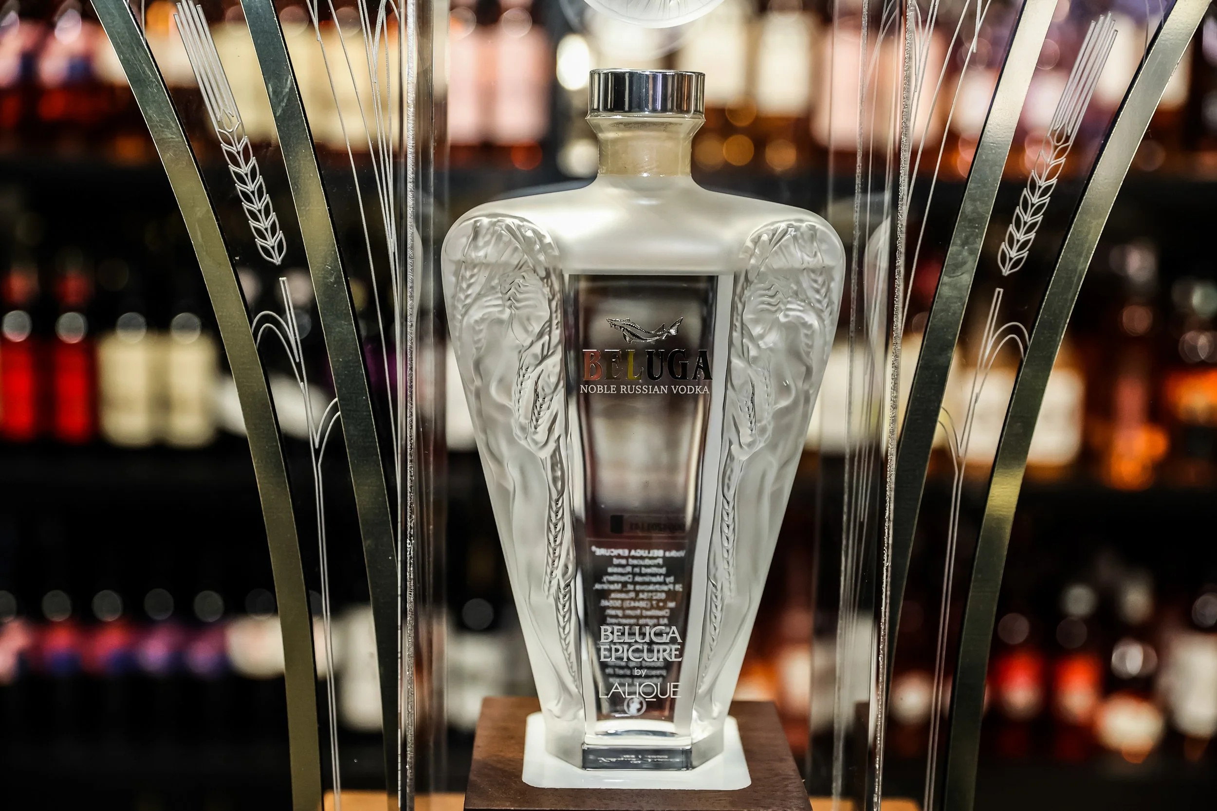 A decorative glass bottle with the label 'Beluga Noble Russian Vodka' on it, displayed inside a metal frame with etched wheat designs. The background shows blurred shelves with bottles.