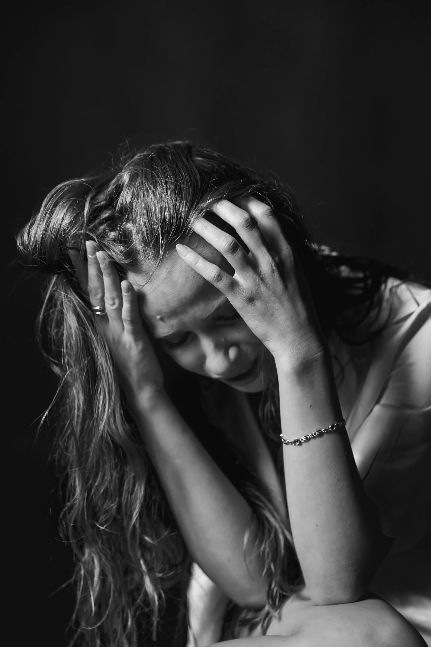 A person with long, wavy hair covering their face, holding their head with both hands, wearing a bracelet on their left wrist, against a dark background.