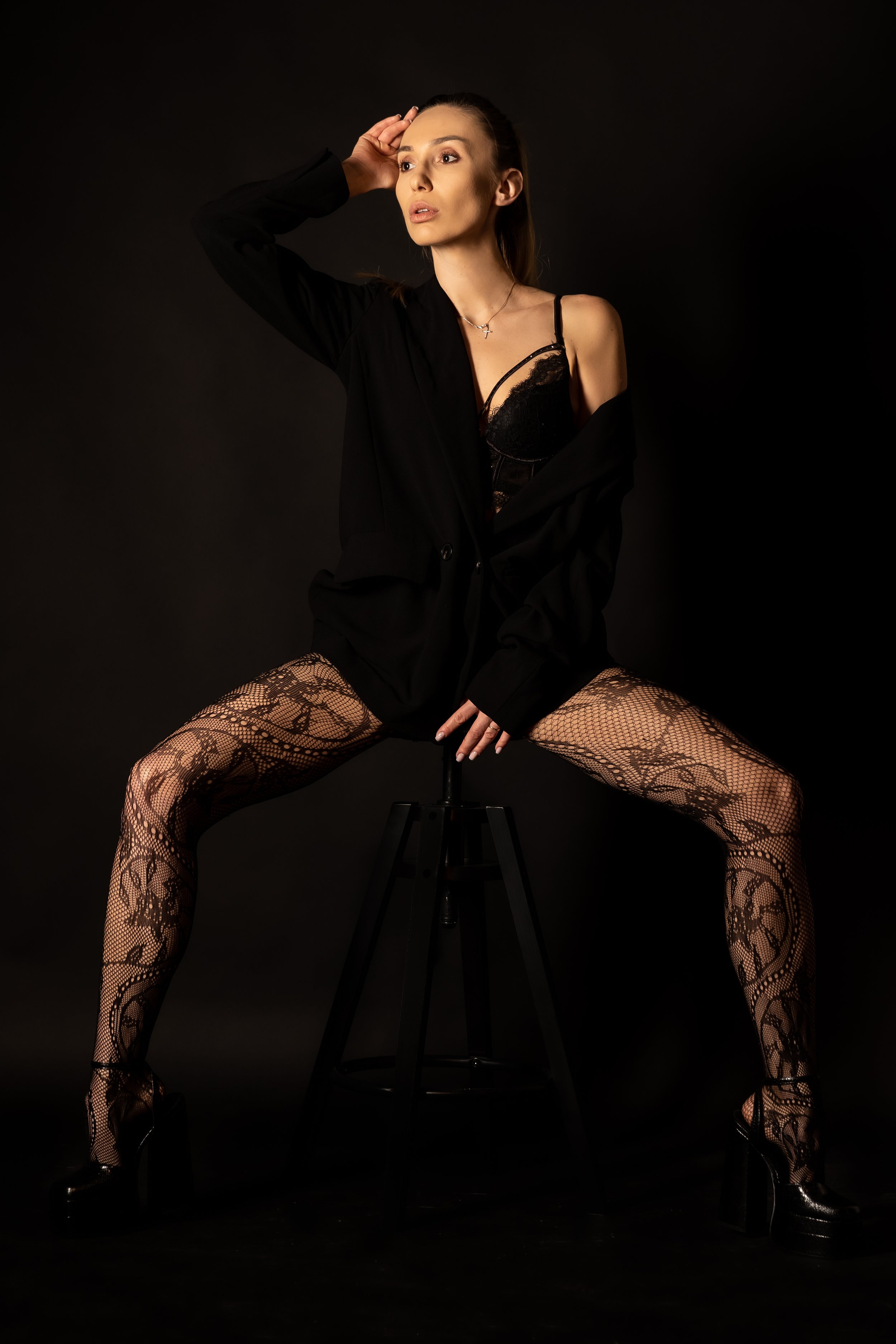 Woman sitting on a stool with legs spread apart, wearing fishnet stockings with floral lace patterns, a black lace bralette, and an open black jacket, posing against a dark background.