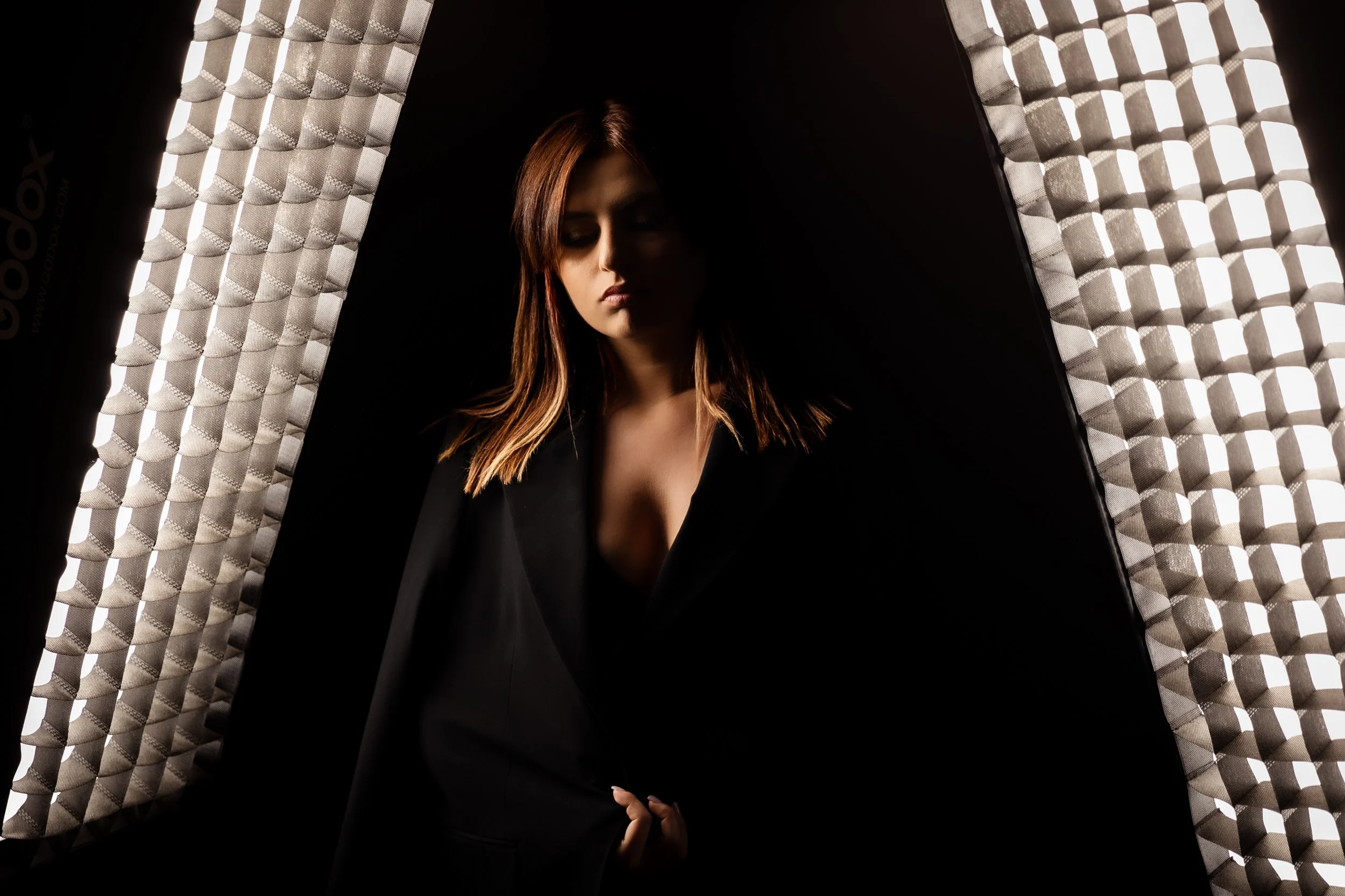 A woman with reddish-brown hair and closed eyes standing between two large softbox lights in a dark room, wearing a black blazer.