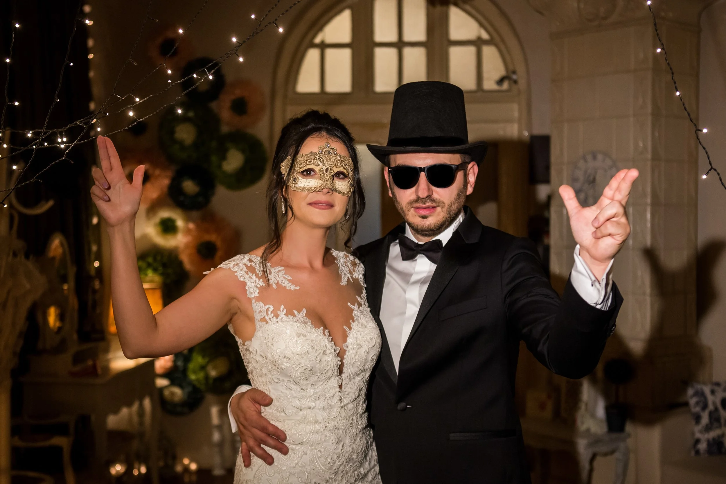 A bride and groom dancing at their wedding celebration, both wearing masks and stylish attire, with festive lighting in the background.