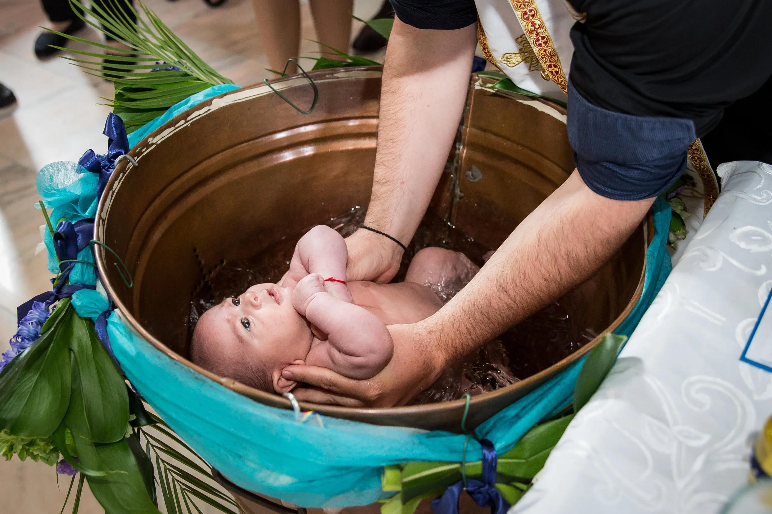 A baby is being baptized in a wooden basin surrounded by green leaves and blue ribbons, with a person gently holding the baby's head.