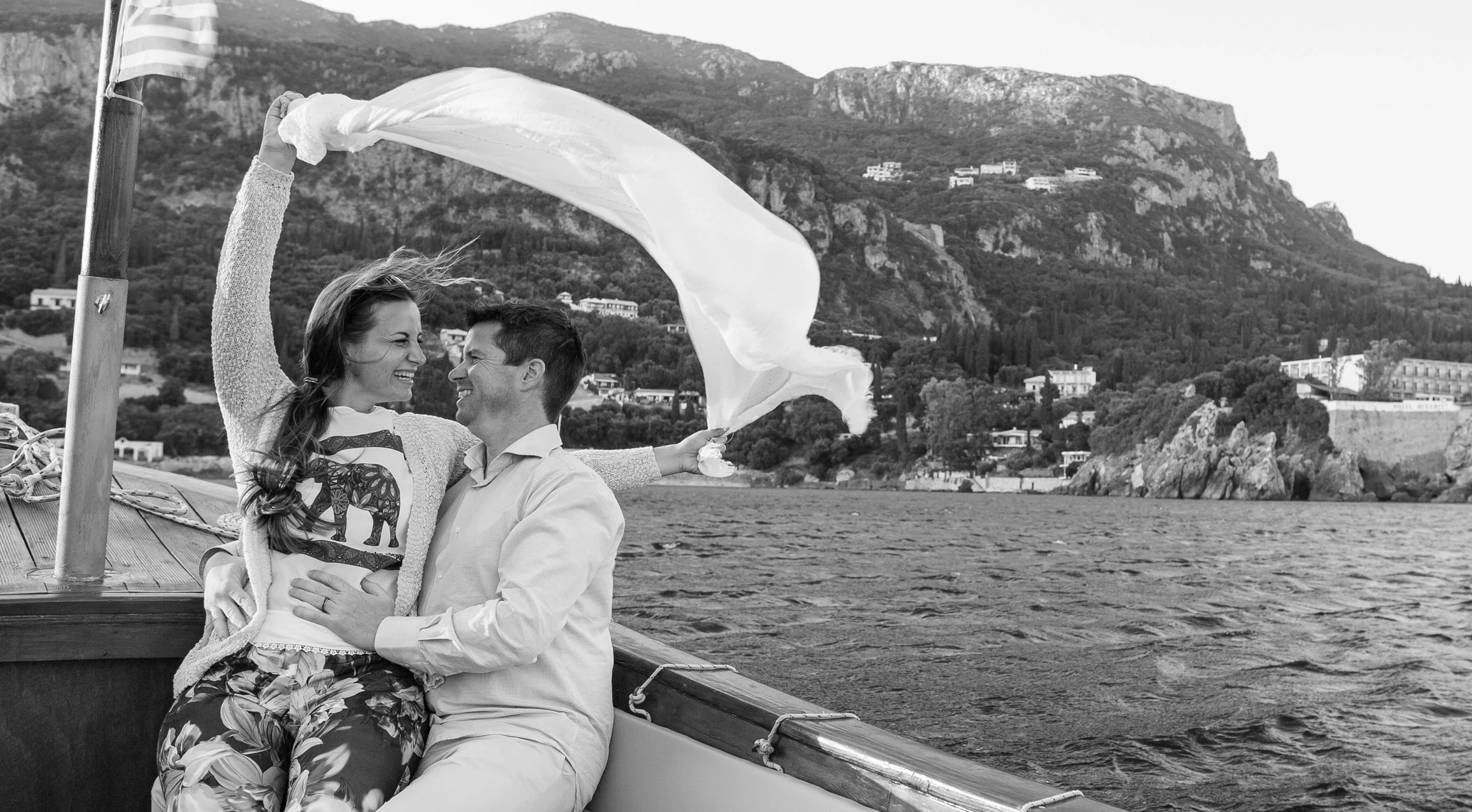 A couple on a boat, smiling and embracing, with a scenic hillside and houses in the background, as they enjoy a moment together on the water.