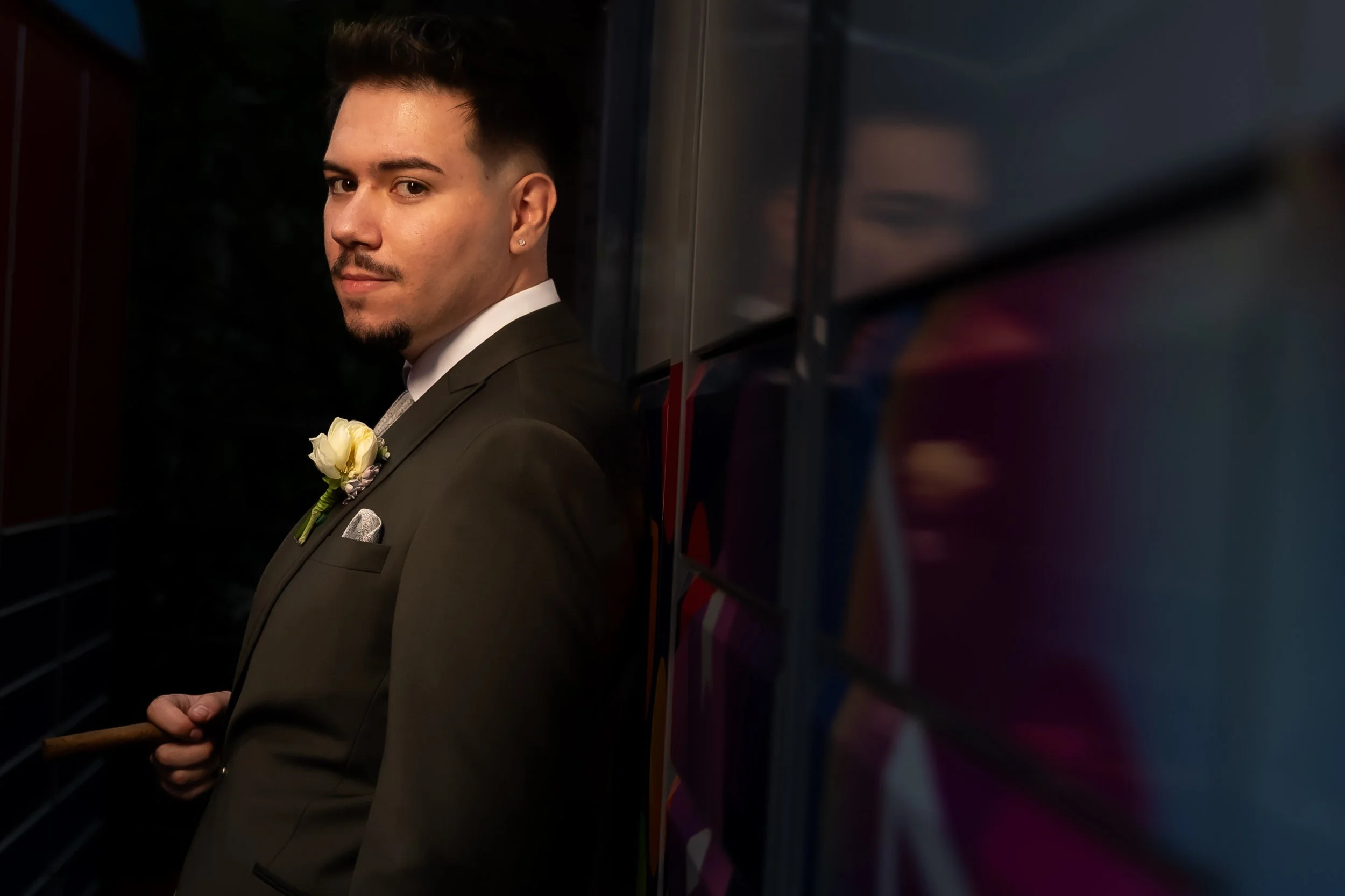 A young man with styled dark hair, light skin, earrings, and a goatee, dressed in a black tuxedo with a white shirt and a tie, standing in front of a reflective surface with colorful patterns, wearing a boutonnière with a white flower.