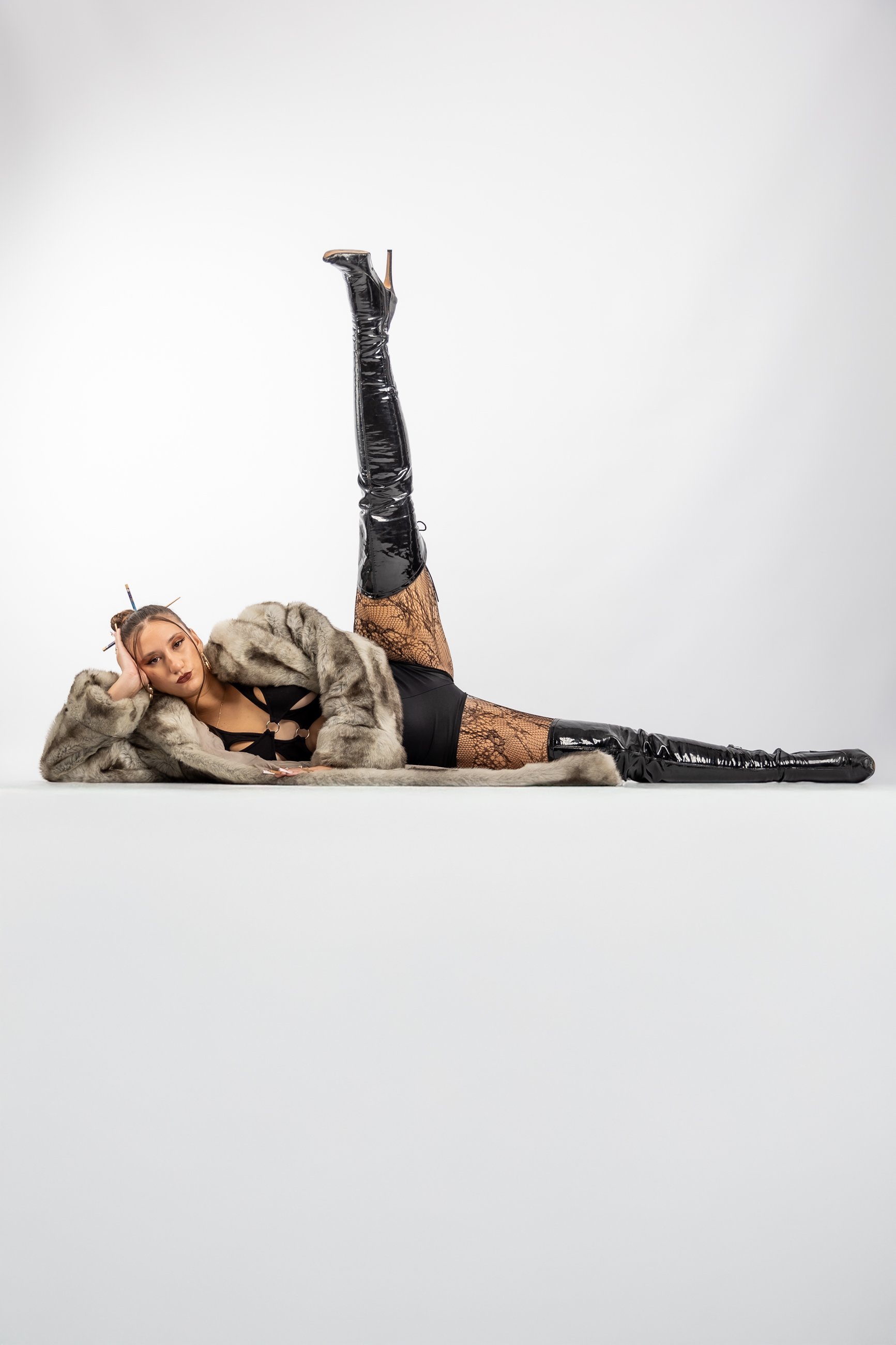 Young woman lying on the floor in a fashion pose, wearing a fur coat, black lace lingerie, thigh-high black patent leather boots, and fishnet stockings, with a plain white background.