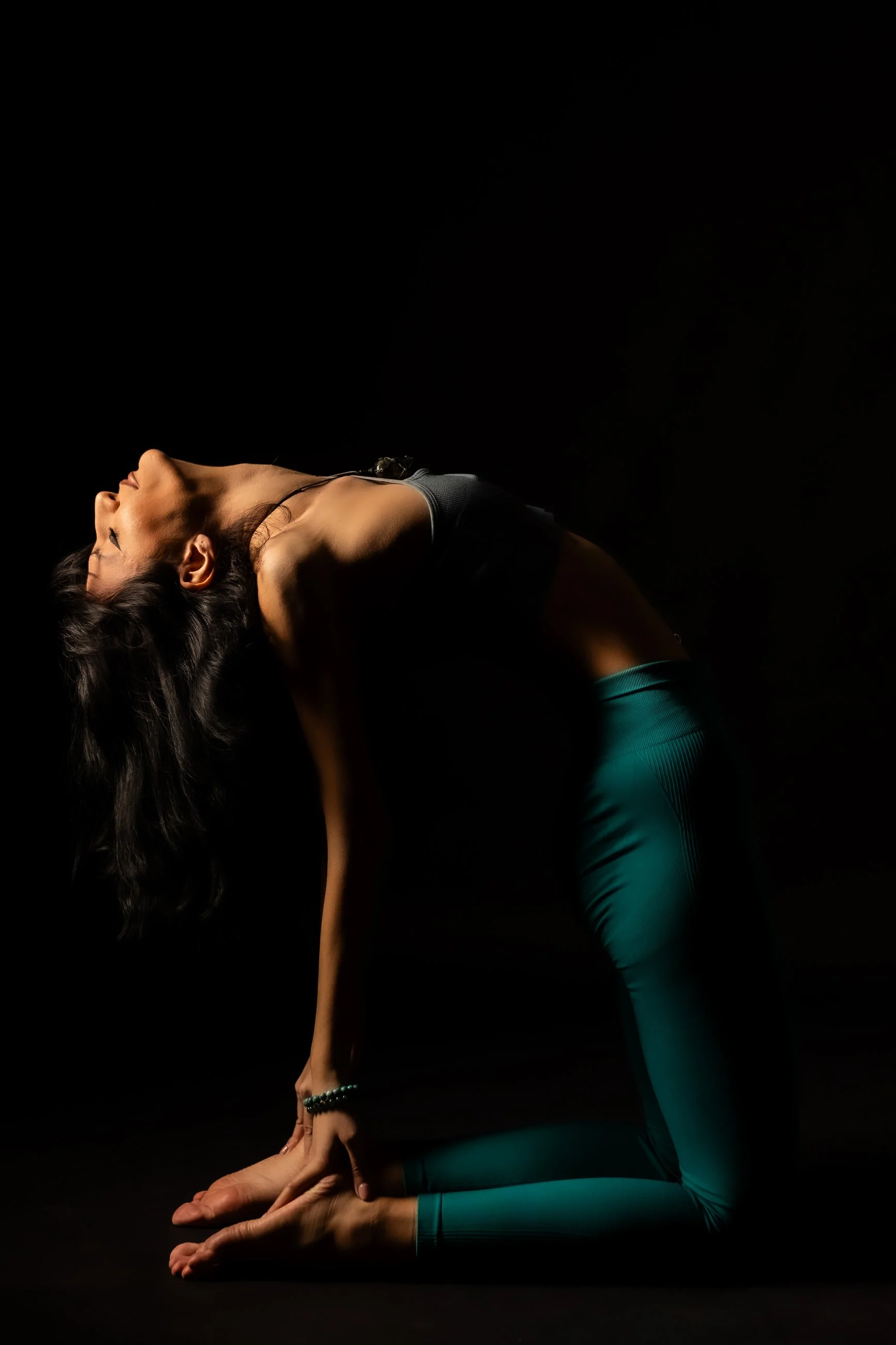A woman practicing yoga, performing a forward bend pose in a dark studio, wearing a black sports top and teal leggings.