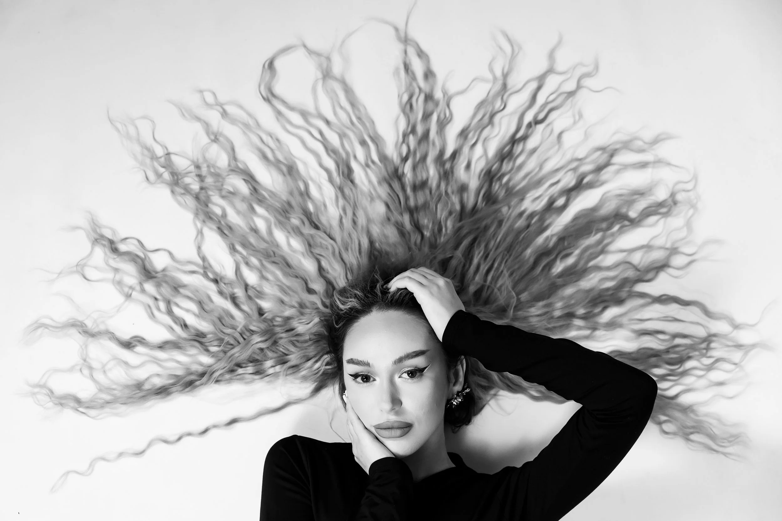 A woman with long curly hair spread out behind her, lying on a white surface, with her head tilted and one hand touching her forehead. She is wearing a black long-sleeve top and makeup with winged eyeliner, looking directly at the camera.