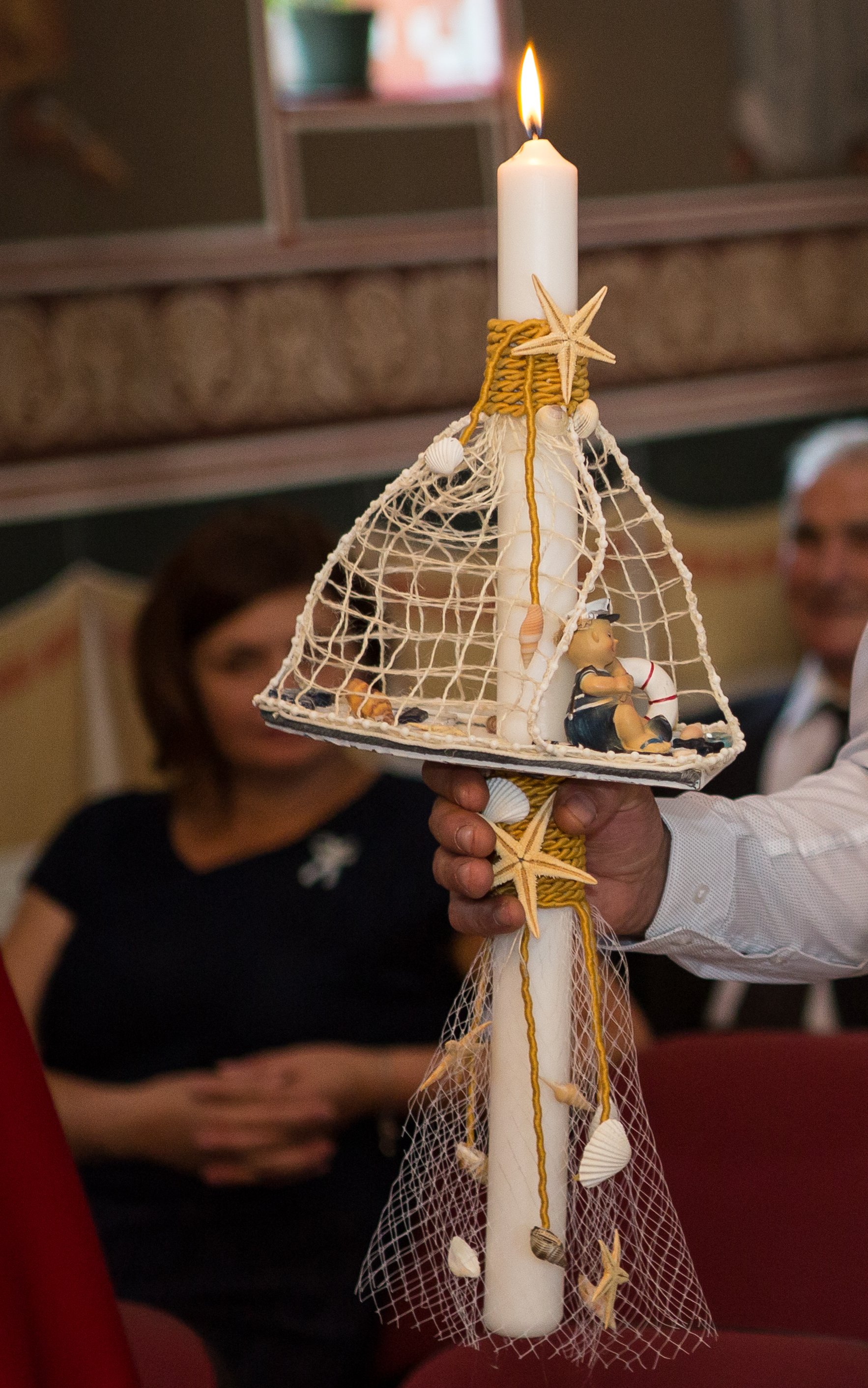 Person holding a candle with decorative seashells and starfish, with people in the background in a room.