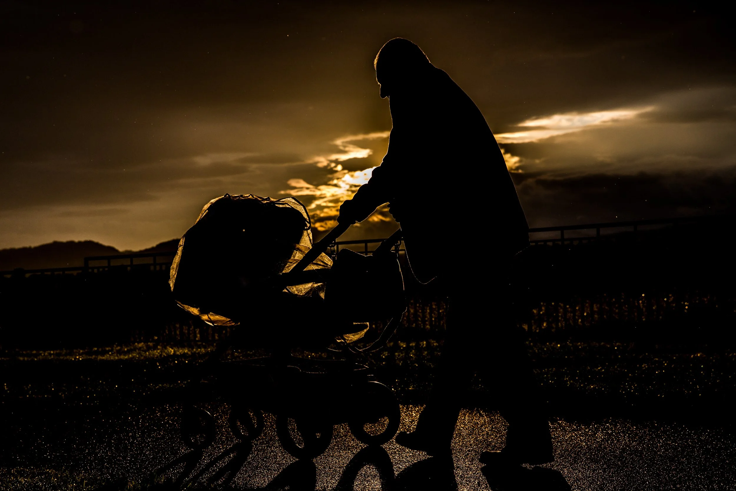 Silhouette of a person pushing a stroller at dusk or night, with a cloudy sky and moonlight in the background, and a fence along the horizon.