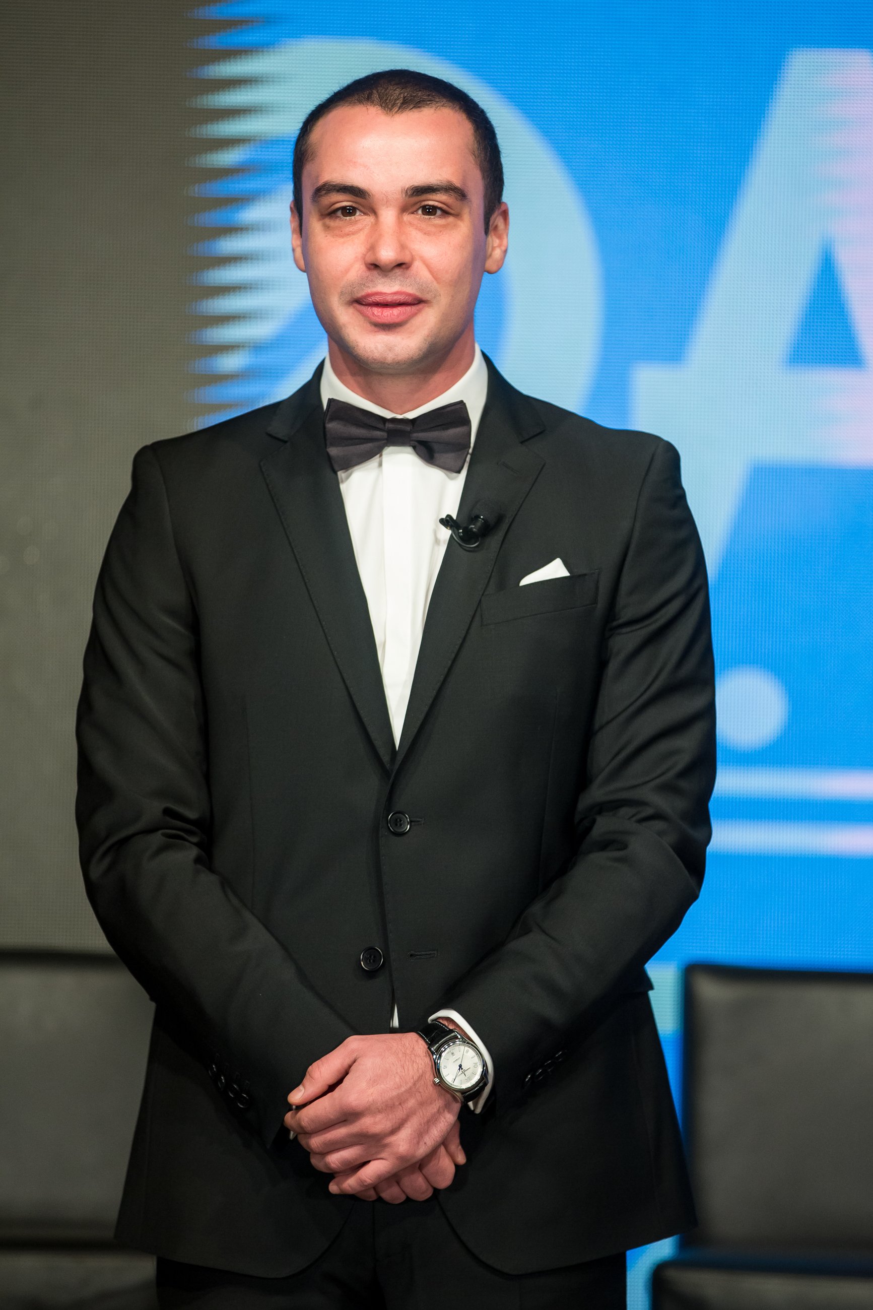A man in a black tuxedo with a bow tie stands in front of a blue background with a logo or graphic. He is wearing a wristwatch and has his hands clasped in front of him.