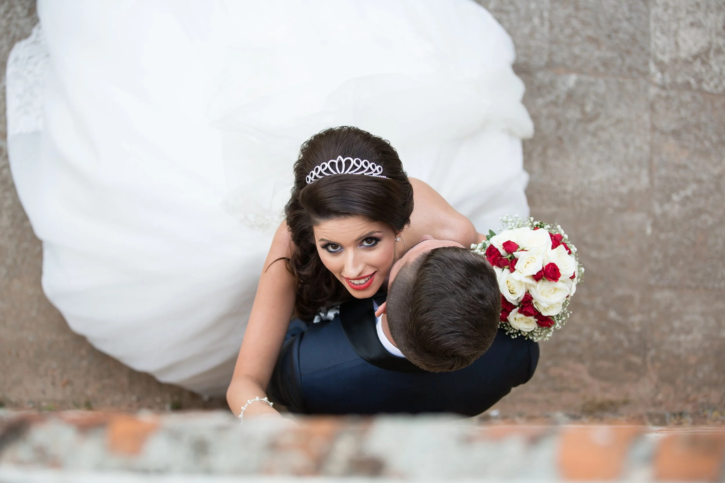 Top view of a bride and groom embracing, with the bride wearing a white wedding dress and tiara, holding a bouquet of red and white roses, against a brick background.