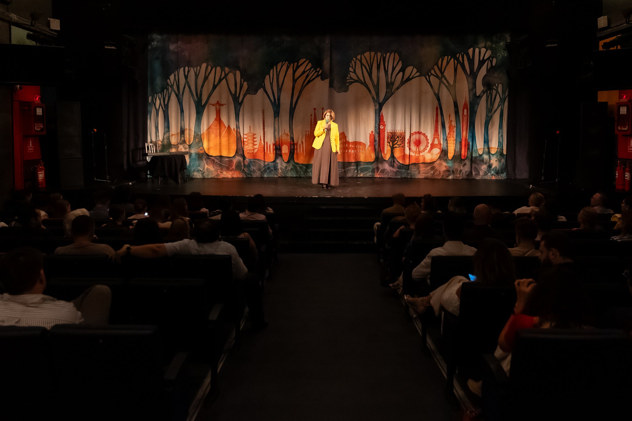 A woman in a yellow blazer speaking on stage at a performance venue with an audience seated in front and a cityscape scenic backdrop.