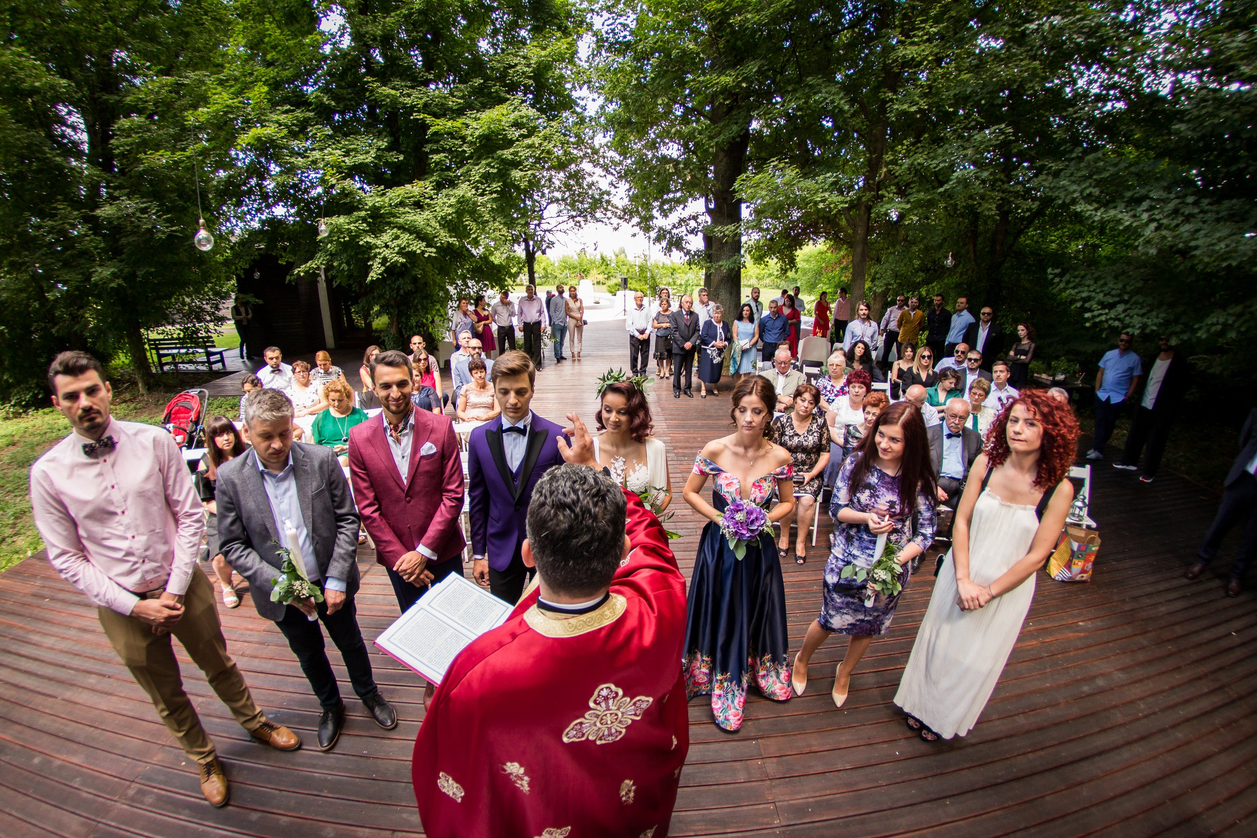 Outdoor wedding ceremony with guests standing on a wooden patio surrounded by large green trees, and a bride and groom facing a person in ceremonial robes.