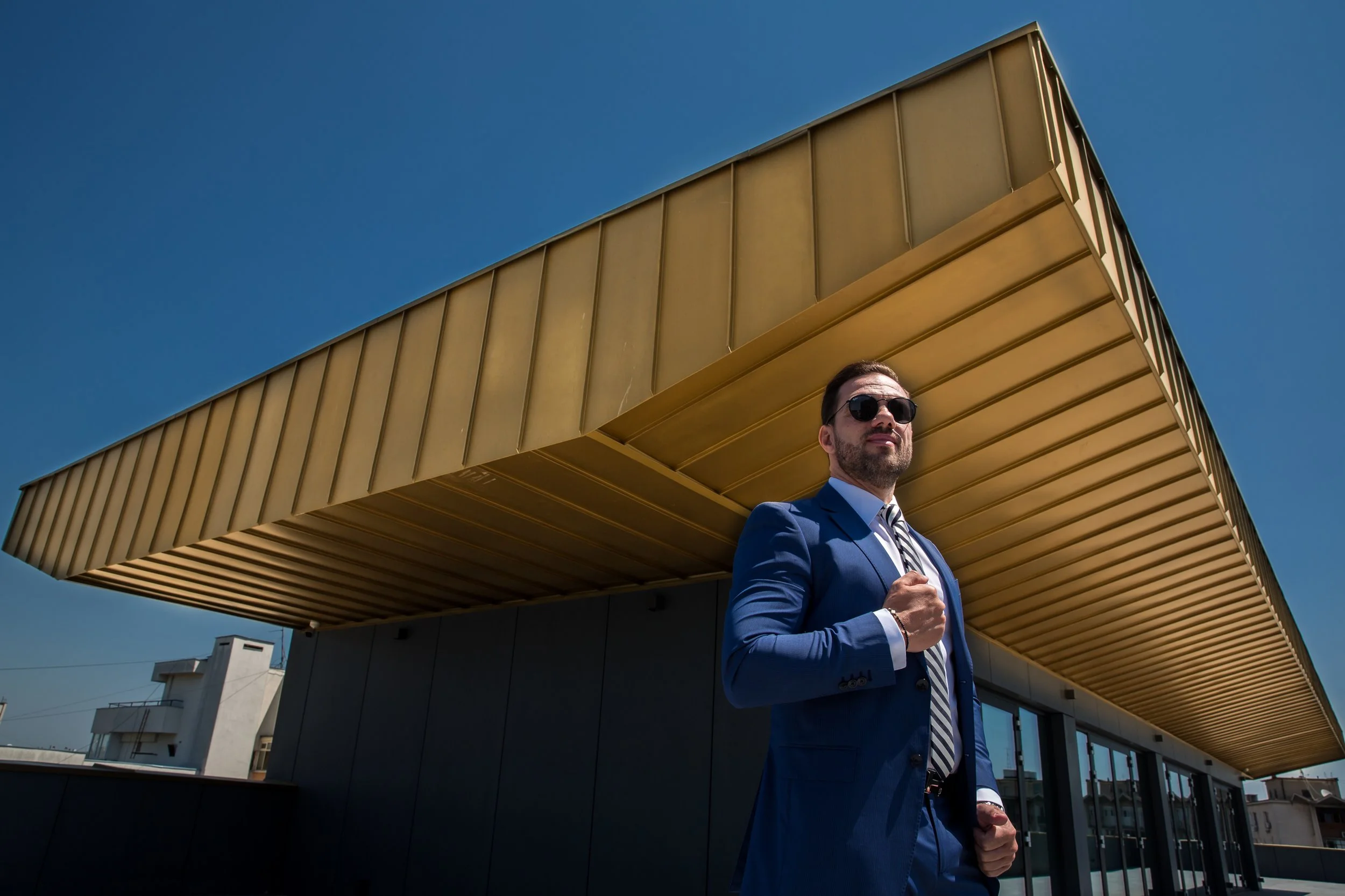 A man in a blue suit and sunglasses walking outdoors under a modern building with a yellow overhang.