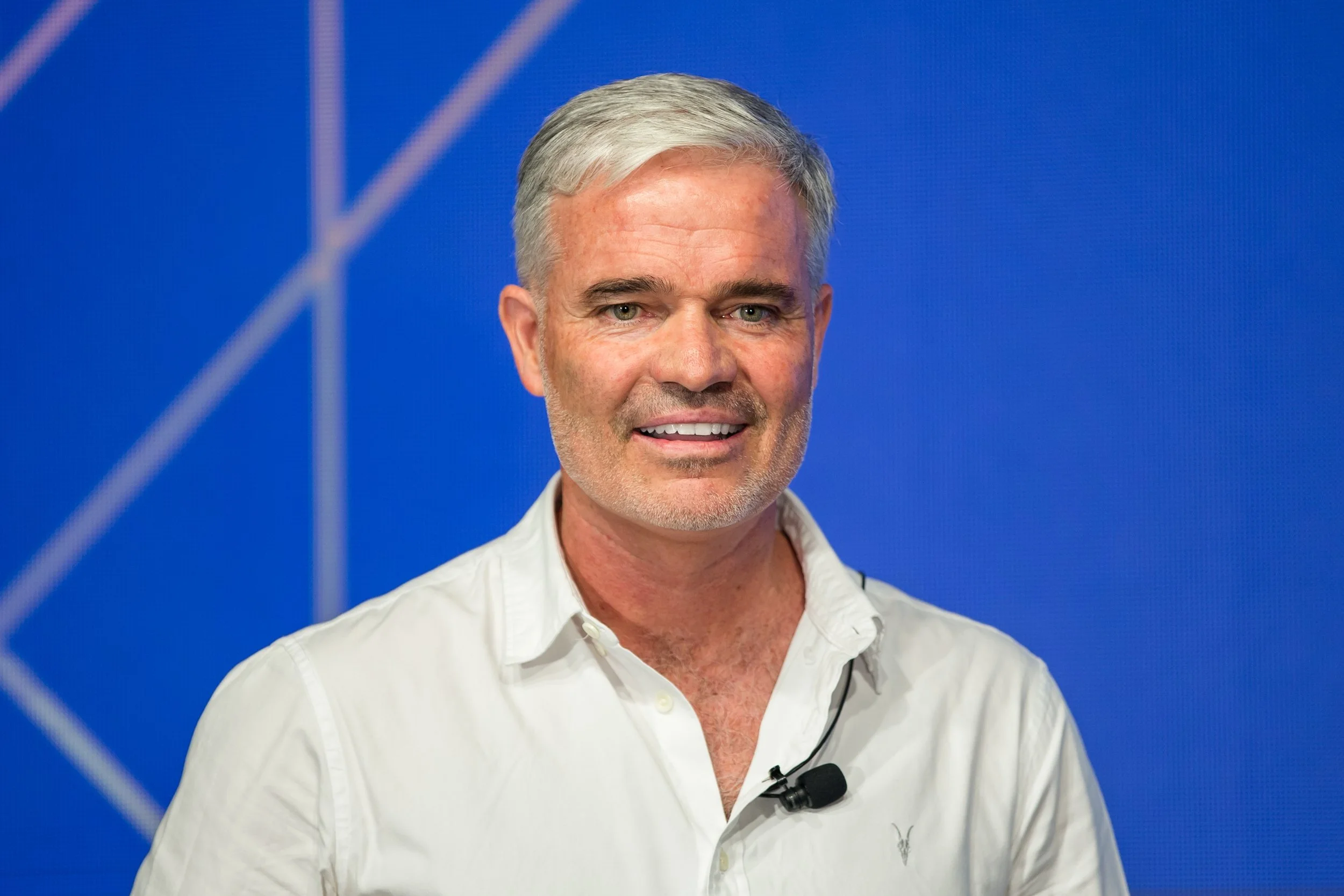 A middle-aged man with silver hair and light skin wearing a white shirt speaking in front of a blue background.