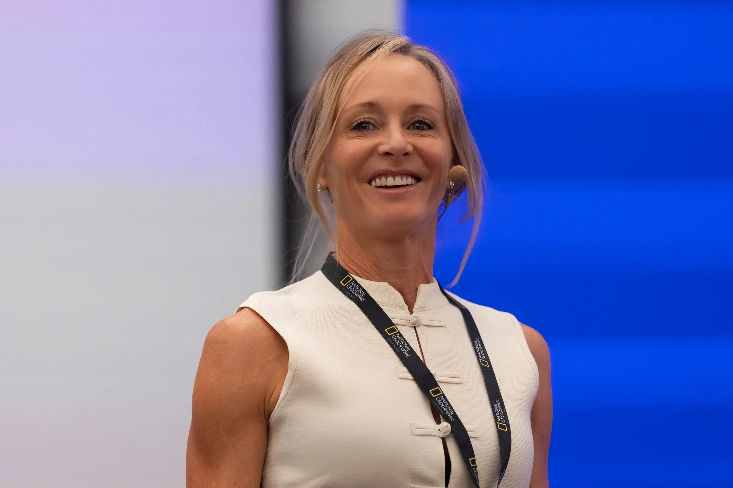 A woman with blonde hair wearing a white sleeveless top and a National Geographic lanyard, smiling and wearing a headset, standing in front of a blue background.