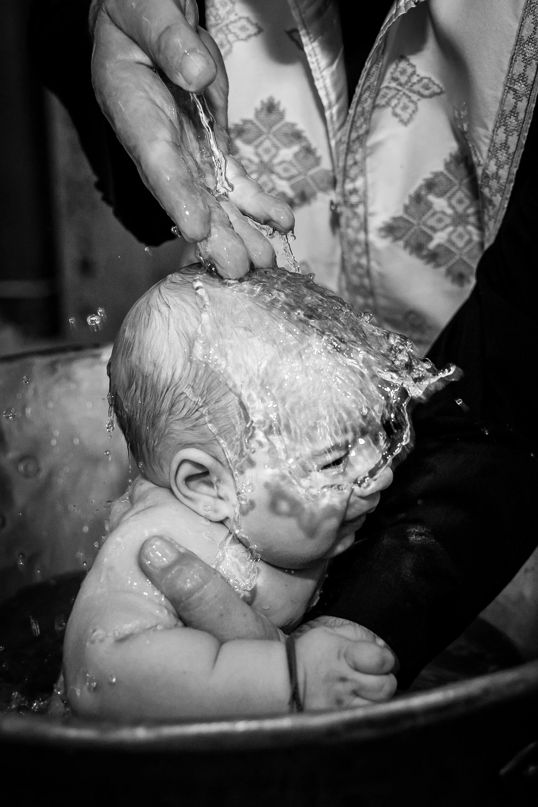 A black and white photo of a baby being bathed, with water splashing on its head and a person holding and supporting the baby's head with one hand. The person appears to be wearing a patterned cloth or apron.