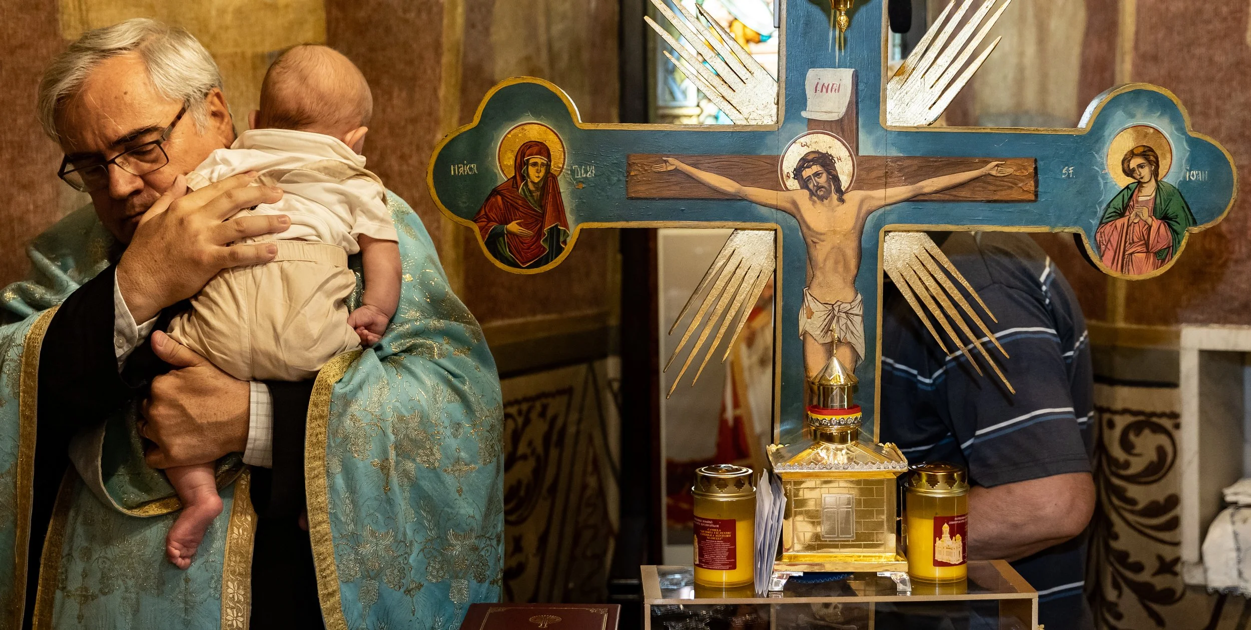 A priest holding a baby during a religious ceremony in a church. There is a large crucifix with icons of saints and candles in front of them.
