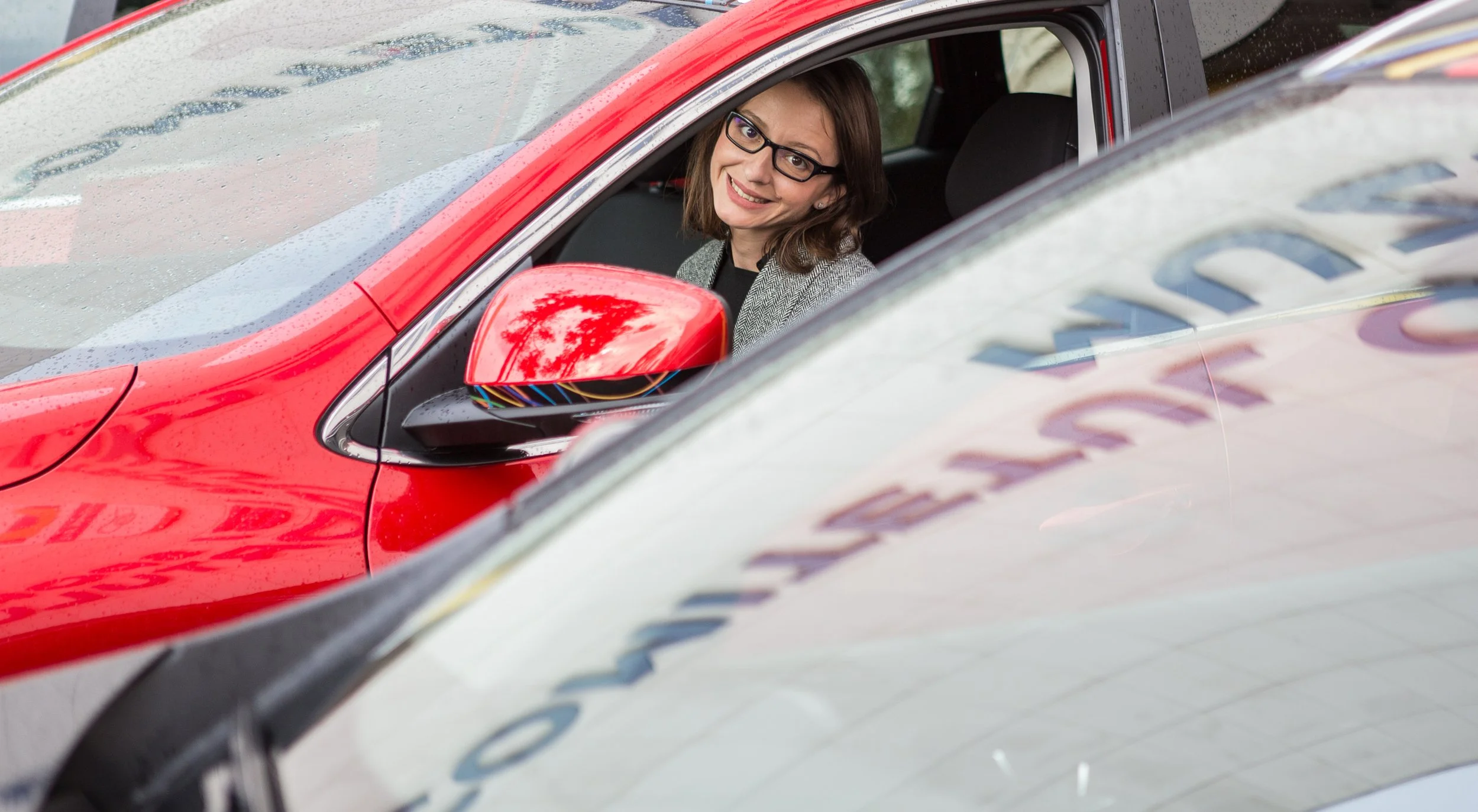 Woman with glasses smiling and sitting in a red car with a rain droplets on the windshield, parked near a police car.